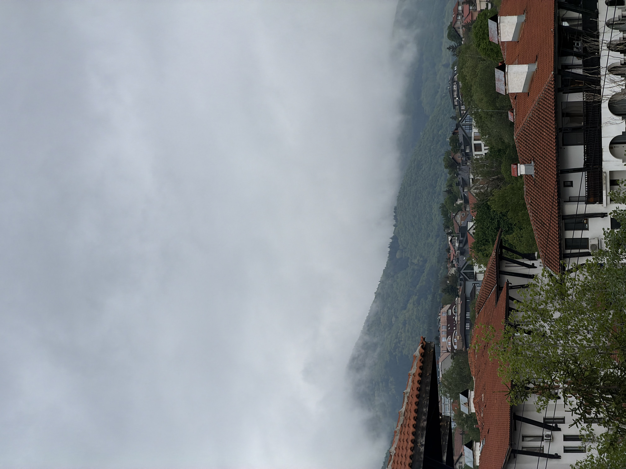 The image depicts a view of a mountain obscured by heavy fog. Adjacent to the mountain, there are buildings with red tiled roofs and white walls, indicative of European architecture. The buildings suggest a residential area, possibly a town or village nestled in the mountains. The dense fog obscures the lower portion of the mountain, creating an atmospheric and somewhat obscured view of the natural landscape. The presence of the buildings indicates a human presence in this location. It is likely located in the city of Sofia, Bulgaria.