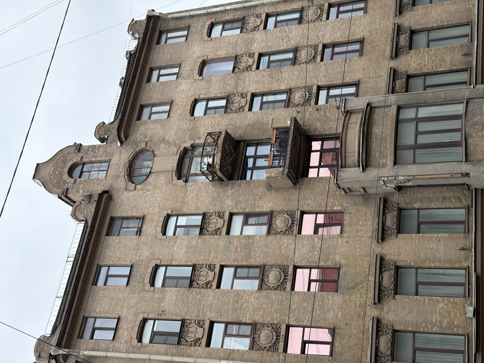 The photograph captures a section of a tall, brick building in Saint Petersburg, Russia. The structure has an elaborate façade featuring decorative elements around the windows and a distinct architectural flair on the left side. Some windows reflect a pink hue, suggesting either tinted glass or a reflection of an adjacent building. Balconies protrude from the central portion of the building, adding depth to the façade. Wires are strung across the top of the frame. The overall impression is of a well-preserved historic building, demonstrating the rich architectural heritage of Saint Petersburg.