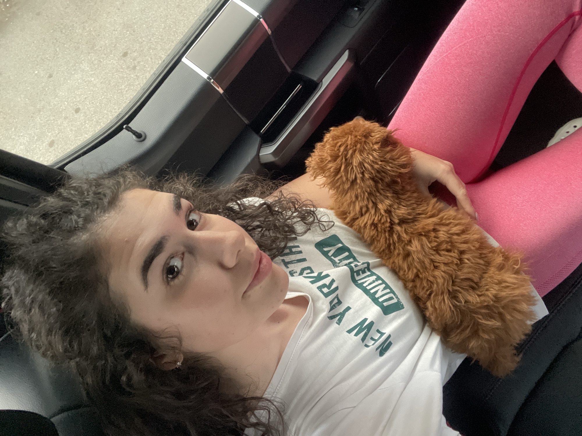 The image shows a young woman and a small, fluffy brown dog inside the passenger side of a car in Sofia, Bulgaria. The woman is reclined, looking directly at the camera with a neutral expression. She has curly dark brown hair, fair skin, and dark eyes. A small, light brown or ginger-colored dog, appearing to be a poodle or similar breed, rests comfortably on her chest, with her right hand gently touching its back.
She is wearing a white T-shirt with the text "NEW YORK UNIVERSITY" printed in dark green letters, visible upside down on her chest. Her lower body is covered in bright pink leggings. The car's interior is dark, with subtle chrome accents visible on the door panel to her left. A door lock pin is clearly seen on the car door frame. Through the open car door, a light-colored ground surface, possibly concrete or asphalt, suggests it is daytime. The scene conveys a relaxed and casual moment during a car journey or while parked.