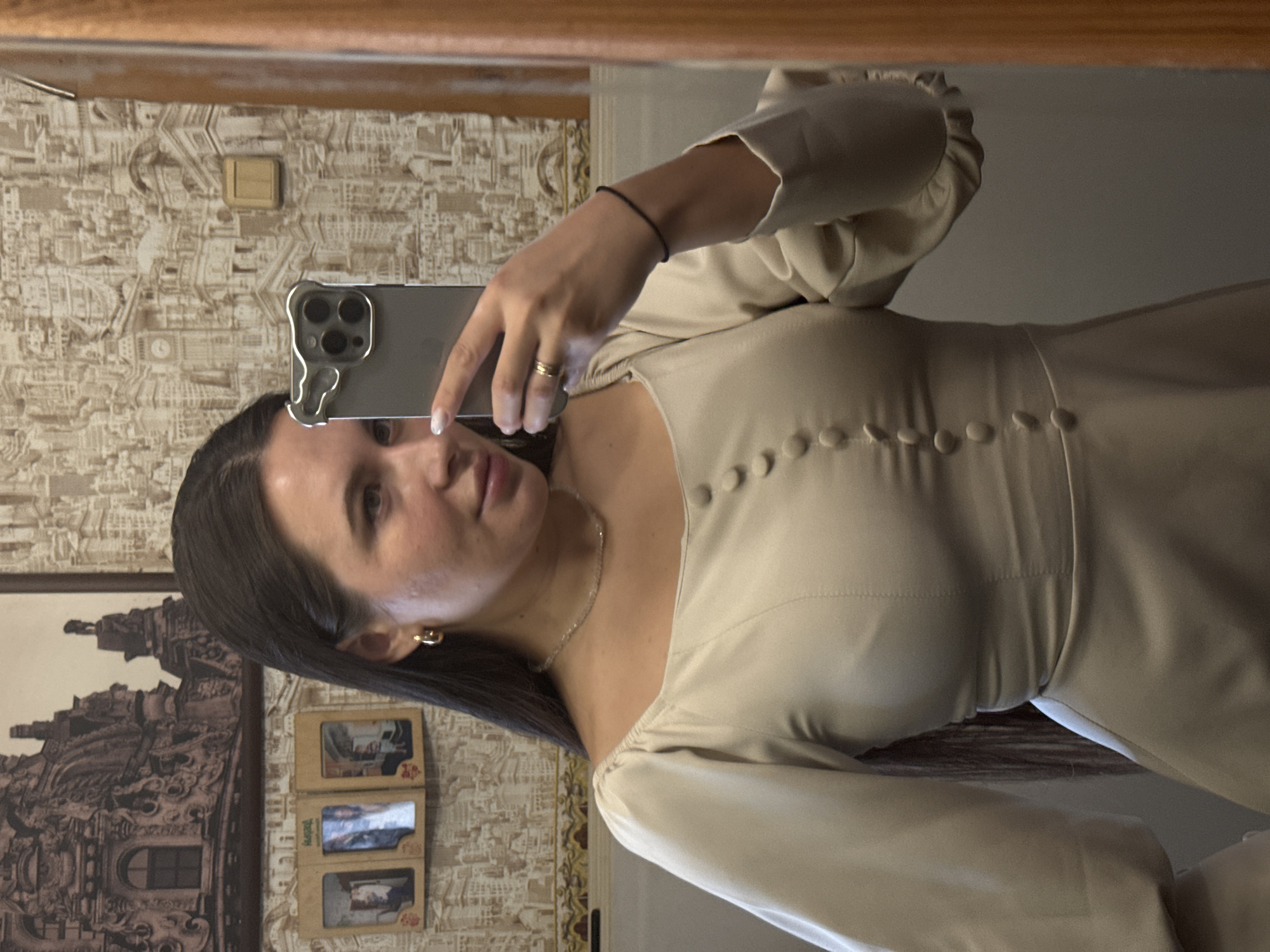 A woman takes a selfie in a mirror. She is wearing a light-colored dress with buttons down the front. She has dark hair and is holding a smartphone. The background includes patterned wallpaper, framed pictures, and a wooden frame. The location is not discernible from the image.