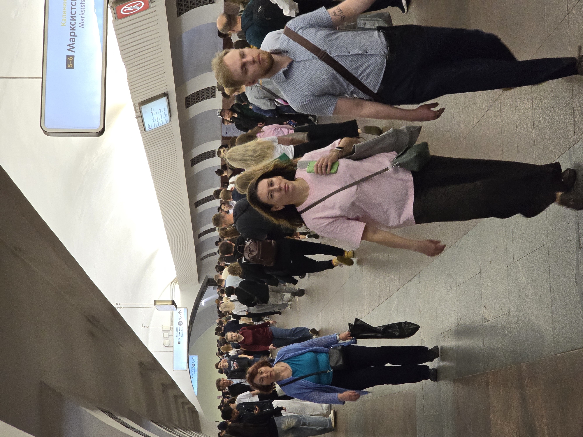 The image shows a crowded hallway in the Moscow Metro in Russia. Passengers are walking in both directions. Signage indicates "Marksistskaya" station. People are dressed in casual clothing.