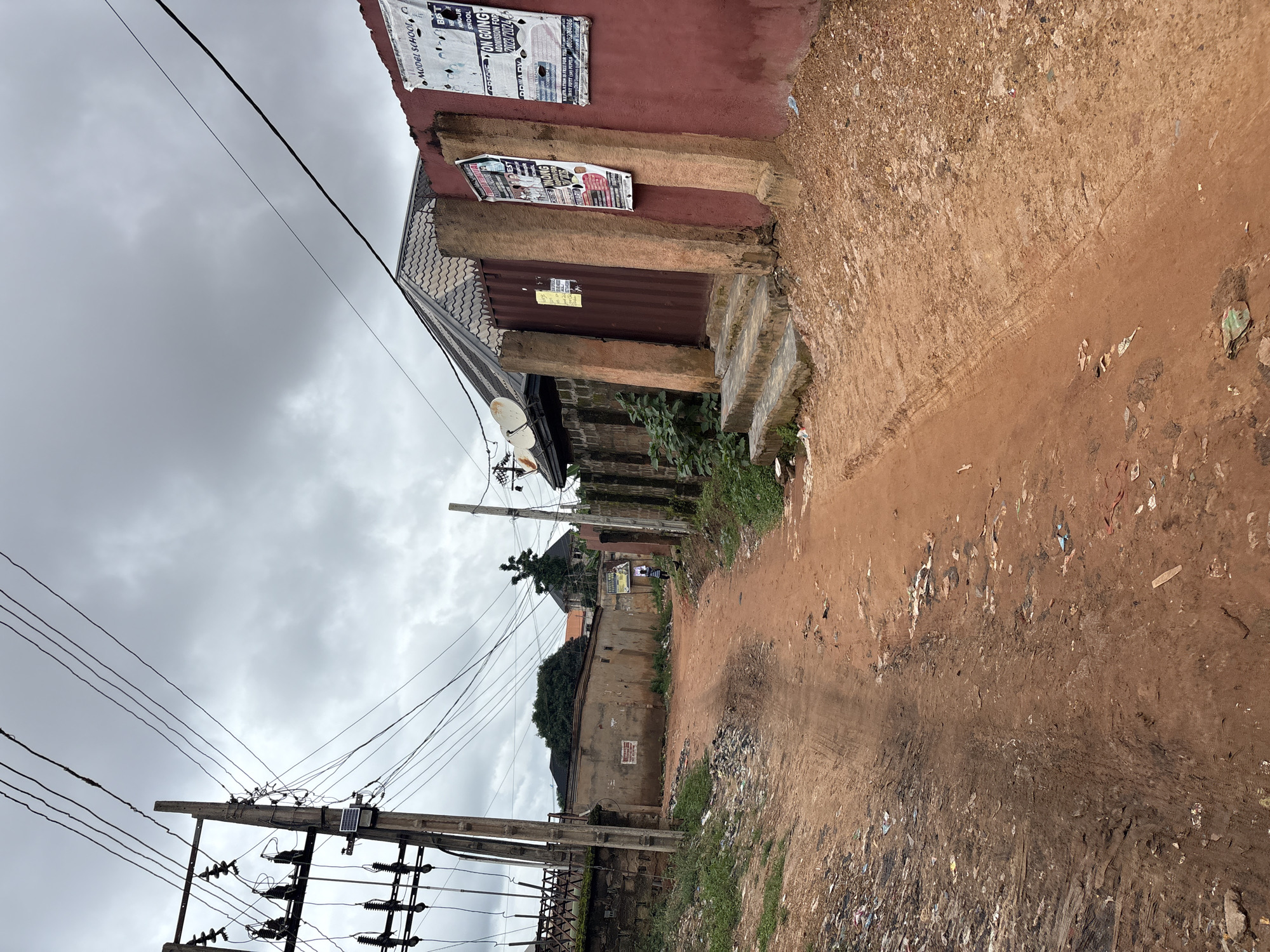The image depicts a dusty, unpaved road in Nkpor, Nigeria. The road is lined with buildings and vegetation. Power lines crisscross the sky, supported by wooden poles. Debris, including plastic waste and scattered papers, is visible along the sides of the road and in ditches. On the right side of the road, a reddish-brown wall is adorned with several posters, some of which appear to be advertisements or announcements. Steps lead up to a metal gate set within this wall. The sky is overcast with gray clouds. A solitary figure, appearing to be a child, is walking away from the camera down the road in the distance. The overall impression is one of a somewhat neglected urban environment.