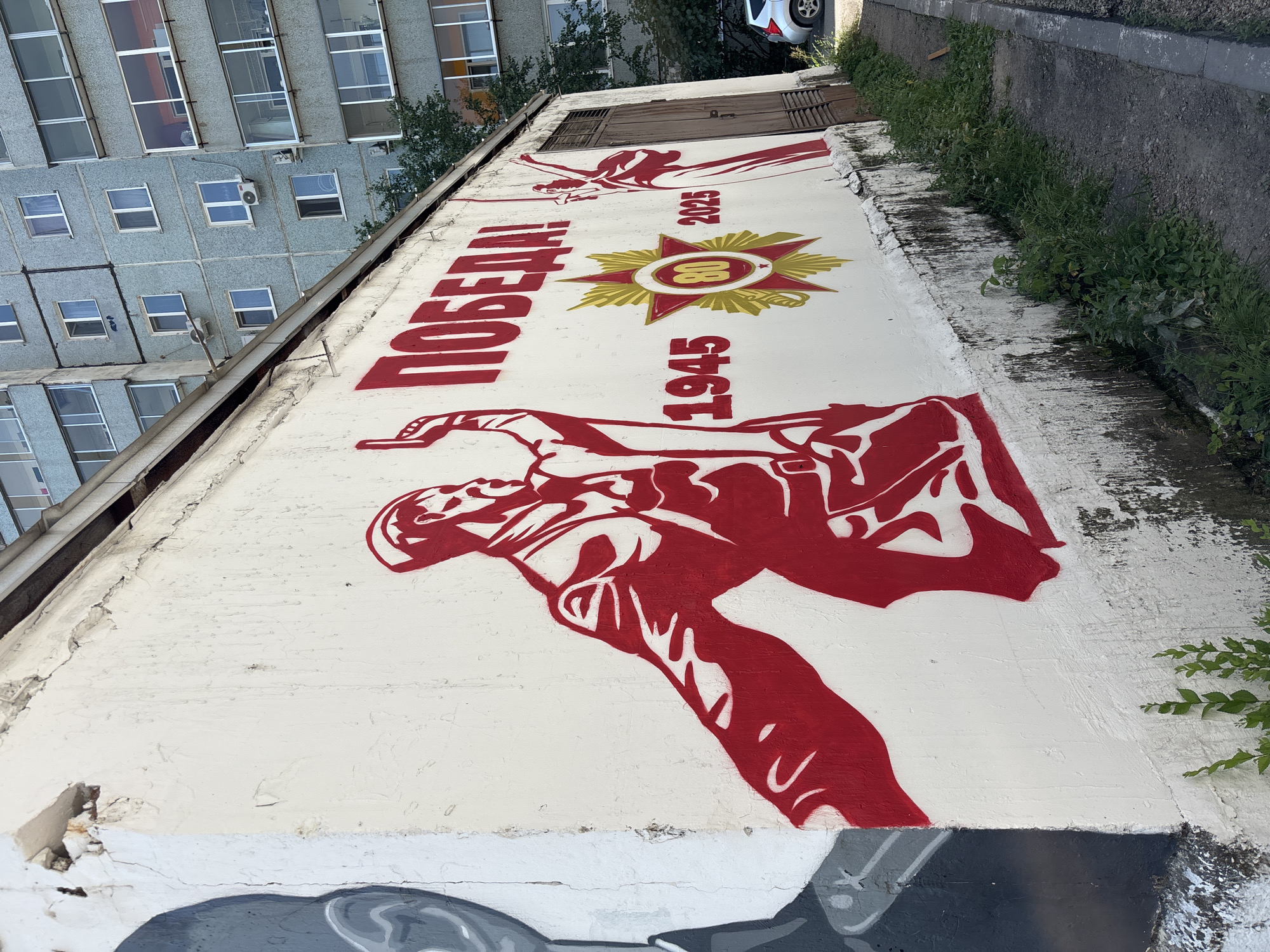 The image shows a mural painted on the side of a building in Blagoveshchensk, Russia. The mural depicts a stylized figure in red, with the word "ПОБЕДА!" (Victory!) painted above it in large red letters. To the right of the figure is a red and gold star emblem with the numbers "80" in the center, flanked by the years "1945" and "2025". The building itself appears to be a low-rise structure with a corrugated metal roof, and in the background, a multi-story residential building with light blue facade and numerous windows is visible. A narrow paved walkway runs along the base of the mural, with weeds and grass growing on either side. A silver car is parked on the right side of the frame. The mural appears to be a commemorative artwork, likely related to Victory Day in Russia.