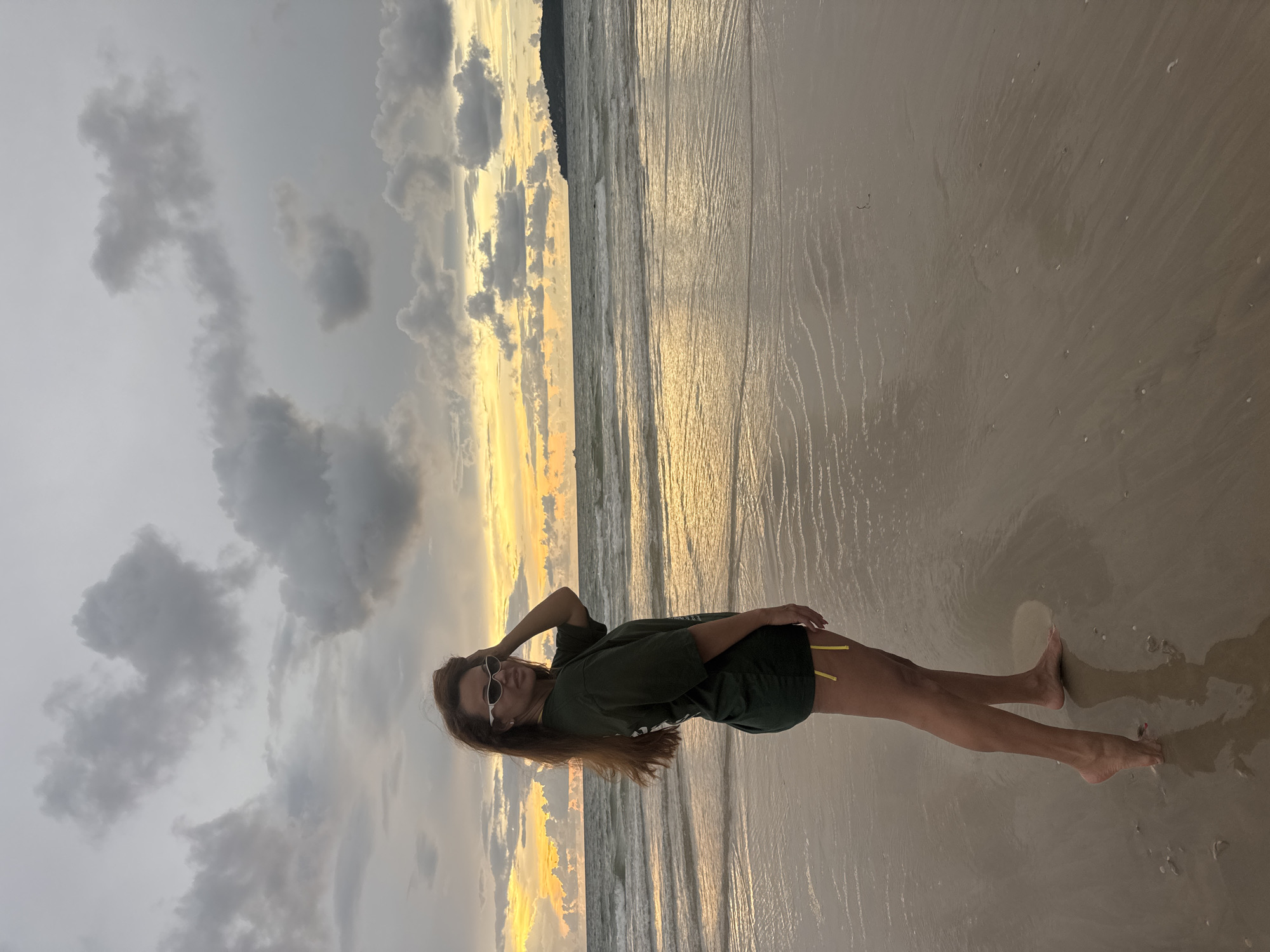 The image depicts a young woman standing on a sandy beach with the ocean and a cloudy sky in the background. The woman is wearing a dark green oversized t-shirt and yellow bikini bottoms. She has long brown hair and is wearing white-rimmed sunglasses. Her left arm is raised, resting her hand on her head. The sand is wet and reflects the light from the setting sun. The sky is filled with dramatic clouds, with streaks of yellow and orange light visible where the sun is setting. The overall scene suggests a serene and perhaps a bit moody atmosphere, typical of a beach at dusk. The location is identified as Choeng Thale, Thailand. There is no visible text in the image.