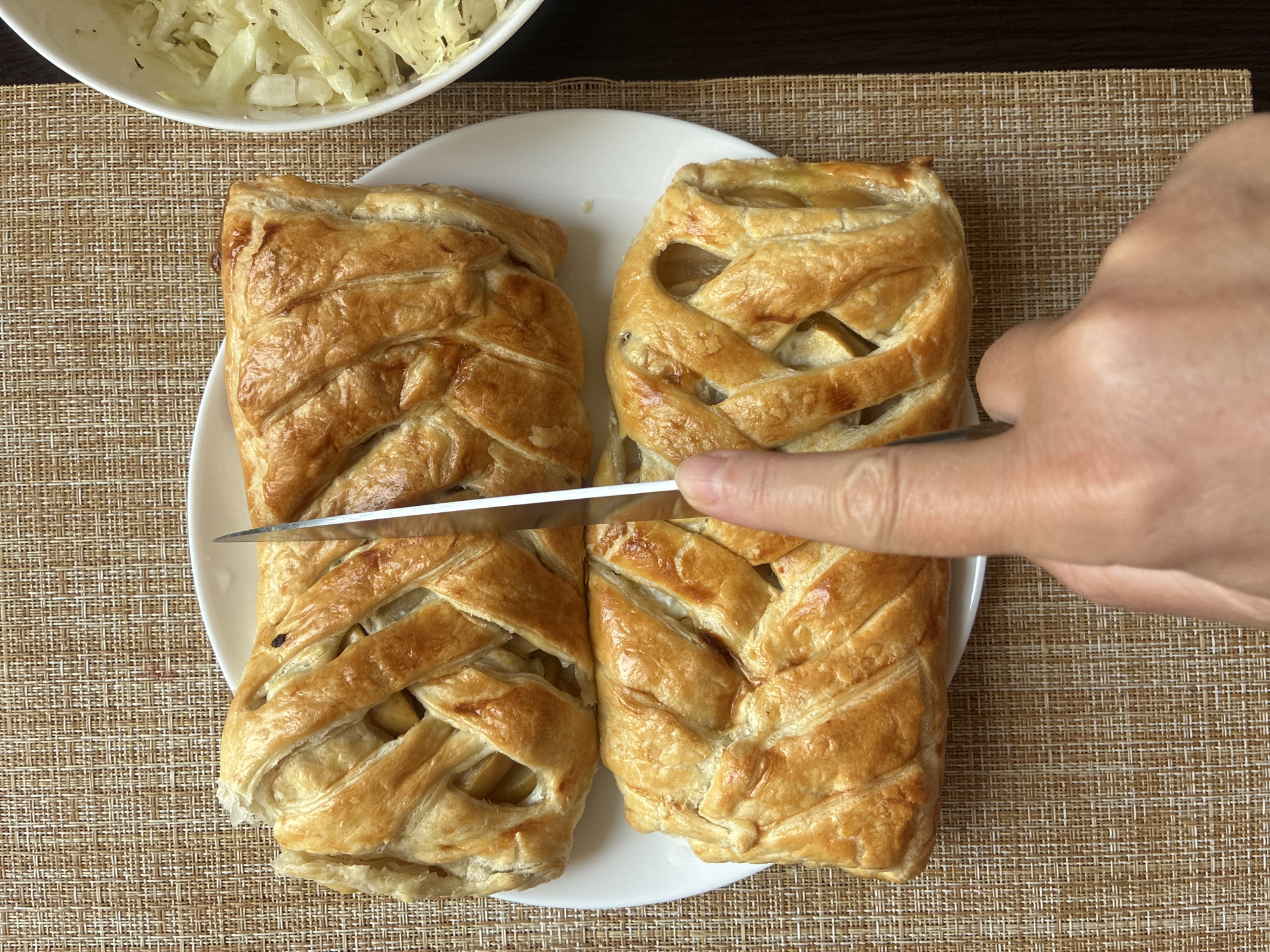 A close-up, top-down shot captures two golden-brown braided pastries on a white plate. A silver knife is positioned in the center, in the process of cutting one of the pastries. A person's hand and finger are visible at the bottom, guiding the knife. To the right of the plate, a white bowl contains a shredded green vegetable, possibly coleslaw, seasoned with black pepper. The scene is set on a woven placemat, indicating an indoor dining environment, possibly in Sochi, Russia, given the mention of the city, though no specific Sochi landmarks are identifiable.