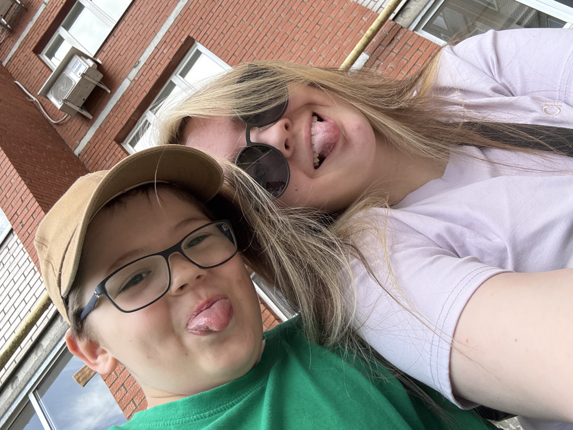 Two people are posing for a selfie outdoors. A young boy wearing glasses and a tan baseball cap is in the foreground, sticking his tongue out. Behind him is a young woman wearing sunglasses and a lavender t-shirt, also sticking her tongue out. Behind them is a red brick building with a window and an air conditioning unit. The location is Chaikovsky, Russia.