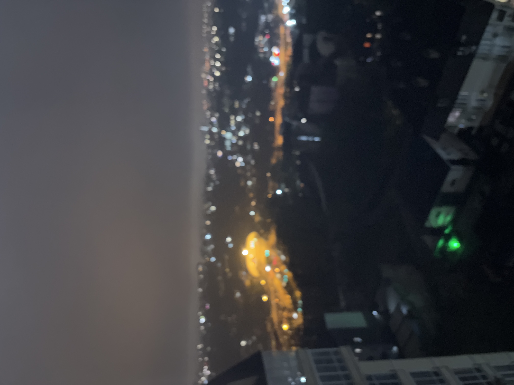The image shows a blurred, nighttime view of a city, likely from a high vantage point within a building. The city lights are visible in the distance, obscured by what appears to be fog or haze. To the right, parts of a building are also visible. Given the prompt indicating the location is Sochi, Russia, it is reasonable to infer that the city lights are those of Sochi, though a definite identification is impossible due to the image quality.