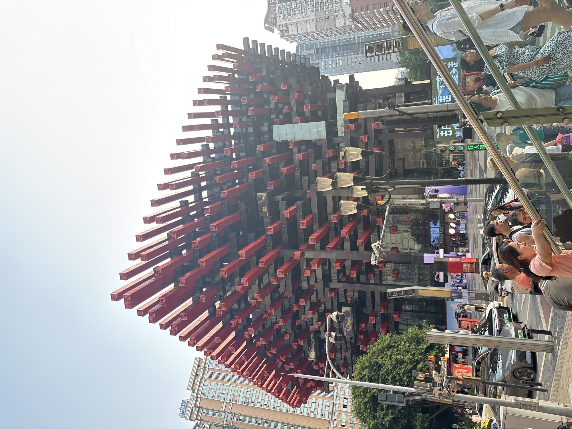The image shows a street scene in Yuzhong District, China, featuring the Raffles City Chongqing horizontal skyscraper. The structure is composed of numerous dark and red horizontal beams. People are walking and riding on public transport on the street below. Cars and traffic signals are also visible. High-rise buildings populate the background. The sky is bright.