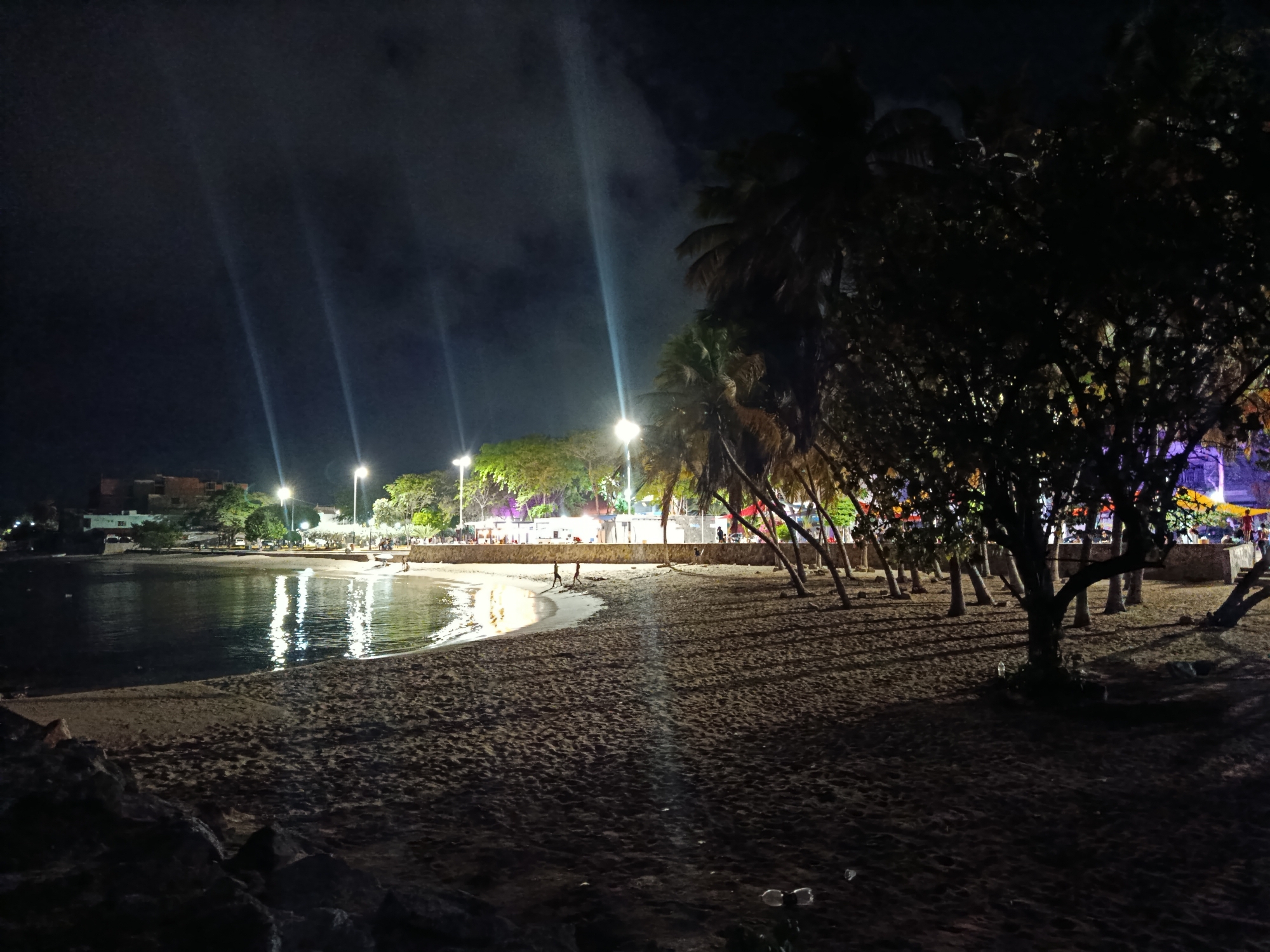 The image depicts a nighttime scene at the beach in Juan Griego, Venezuela. The foreground shows dark sand with some rocks in the lower left corner. The middle ground features a crescent-shaped beach with the calm ocean on the left. The water reflects the lights from the shore, creating shimmering patterns. Several streetlights illuminate the beach area, with their beams cutting through the dark sky. Palm trees line the edge of the beach, casting long shadows on the sand. In the background, buildings and a bustling promenade with colorful lights and a few people are visible. The overall atmosphere is festive and lively, despite the darkness of the night.