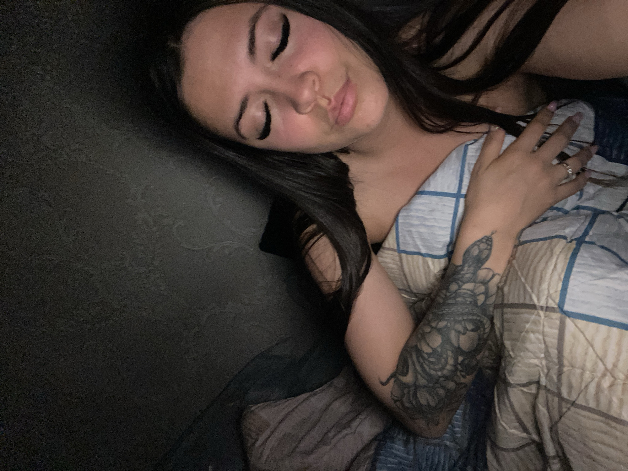 The image depicts a young adult female lying in bed within a dimly lit indoor setting. Her long, dark hair falls around her shoulders and chest. Her eyes are closed, accentuated by long false eyelashes, and her lips are softly pursed. A prominent tattoo of a snake and floral patterns covers her right forearm, which is visible above a plaid blanket featuring blue, beige, and white squares. Her left hand, adorned with a ring on the fourth finger, rests on her chest, partially obscured by her hair. A striped pillow and a dark, textured wall are visible in the background. The visual content of the image does not provide verifiable details to confirm a specific location in the city of Chaikovsky, Russia.