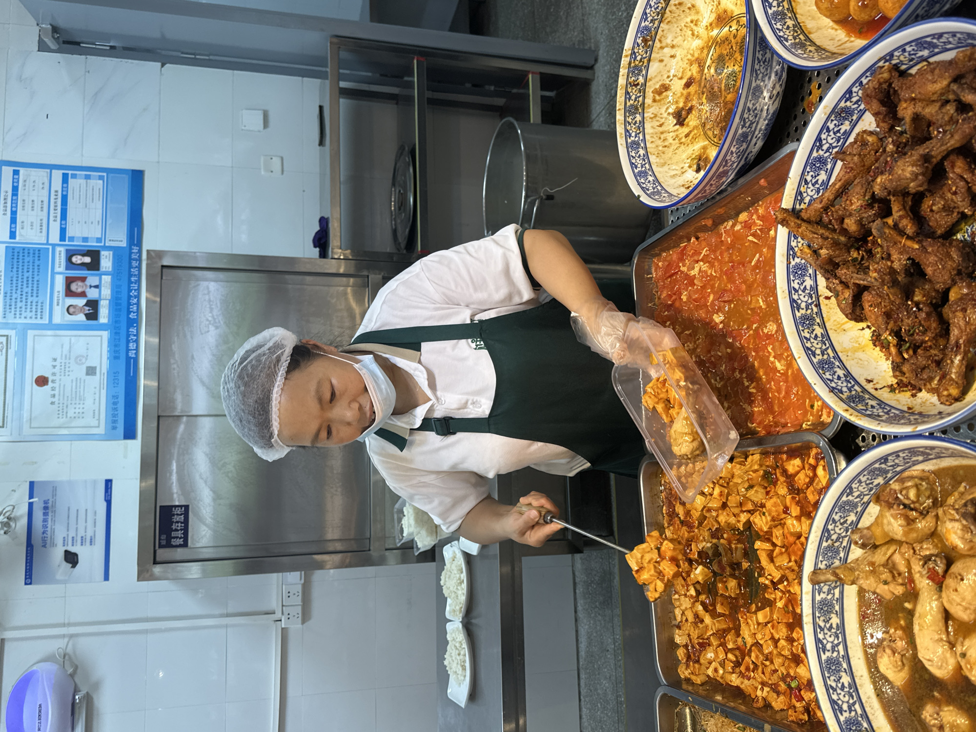 A person wearing a hairnet, mask, white shirt, and green apron is working in a food service environment. They are using a long-handled utensil to serve food into a clear plastic container. The food is in a metal tray alongside several platters of food, including tofu and chicken. Behind the person are stainless steel surfaces. On the wall, there are two posters and a sign that says "Food Catering License." The lower text on the poster mentions Jiangjin District Market Supervision Bureau, Chongqing City with the phone number 47519300. The second sign mentions a "AI action identification camera". A third sign mentions "Tableware storage cupboard".