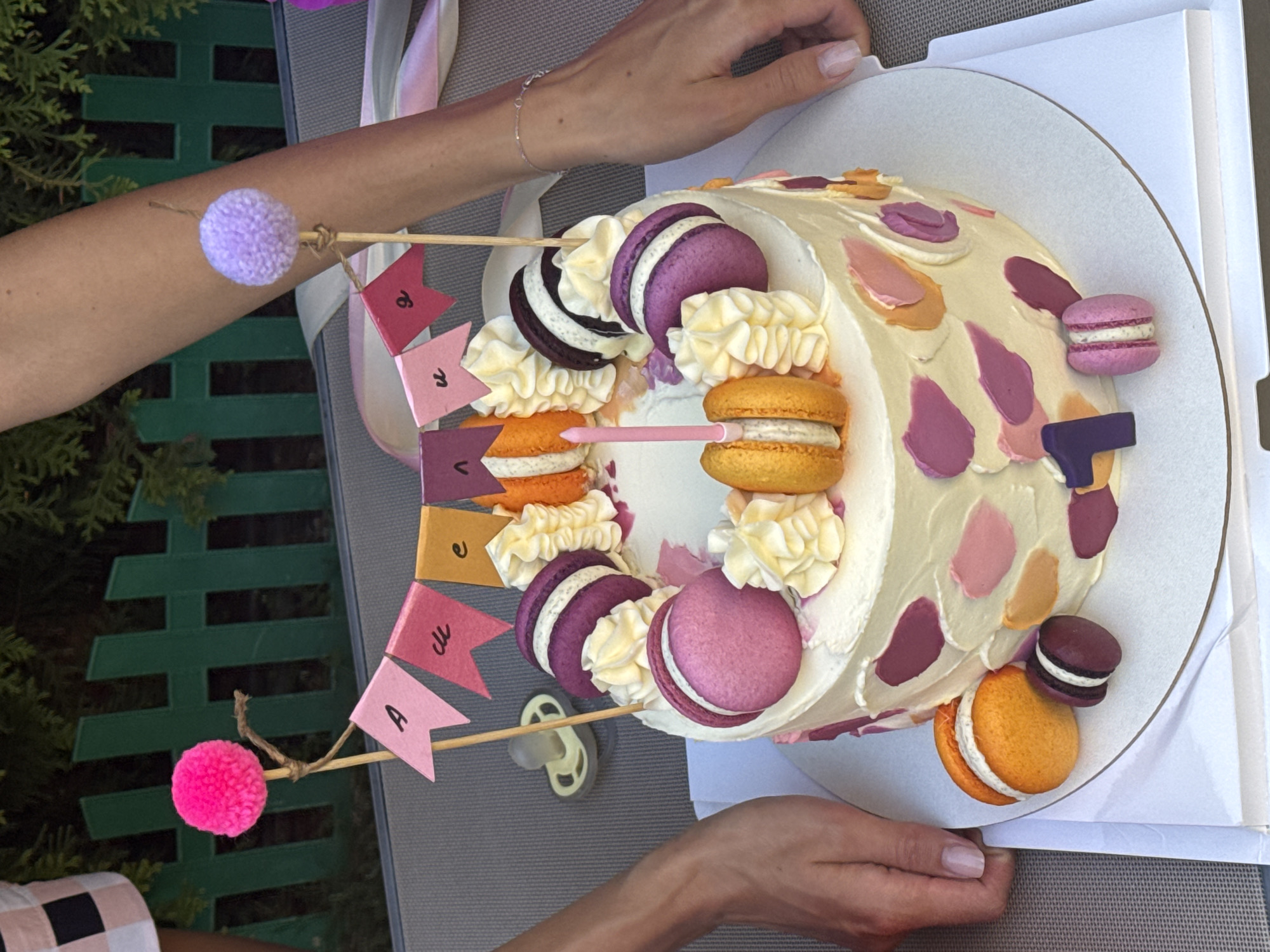 The image shows a white birthday cake decorated with macarons, piped frosting, and a banner that reads "Alice". The cake has colorful frosting splotches on its sides. A single pink candle is present. On the left side of the cake, a pacifier is visible. The cake is placed on a table outdoors, with a green fence and greenery in the background. Two women's hands are visible, one on the left and one on the right, as if they are presenting or interacting with the cake. The scene appears to be in a backyard or garden setting. The location of Sary-Oy, Kyrgyzstan is not depicted in this image.