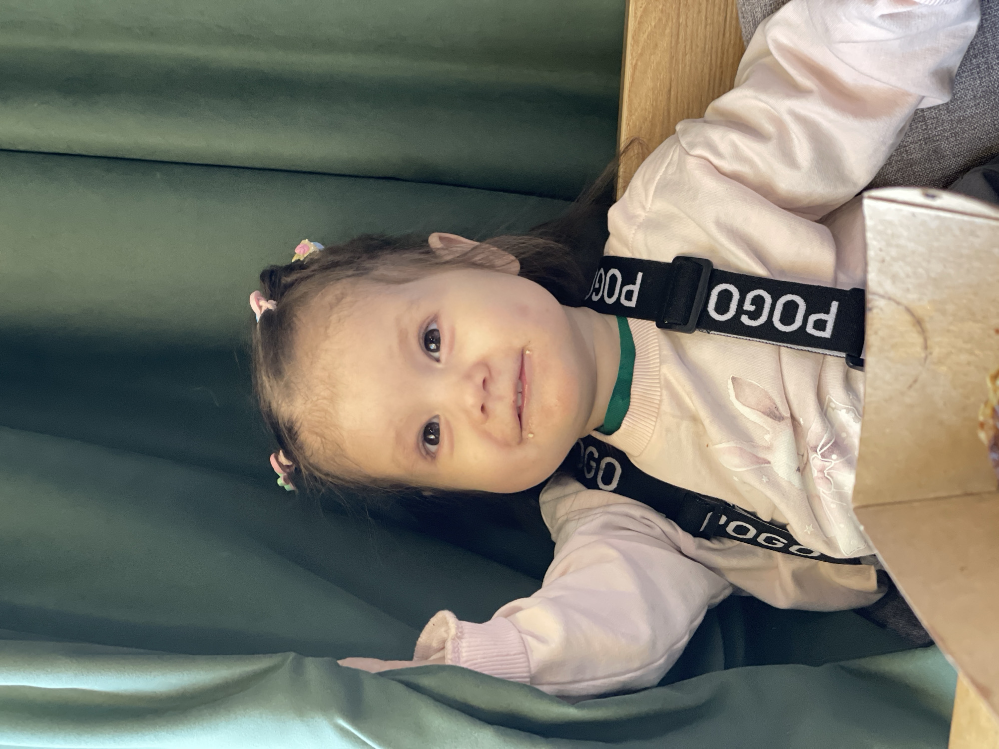 A young child is seated, secured by black straps with white "POGO" lettering, in an interior setting. The child wears a light pink, long-sleeved shirt and has small, colorful clips in their hair. There is some light-colored residue around the child's mouth. Green fabric is visible behind and beside the child, possibly part of the seating arrangement. A wooden structure is adjacent to the child. The specific location within Izhevsk, Russia, cannot be determined from the image alone.
