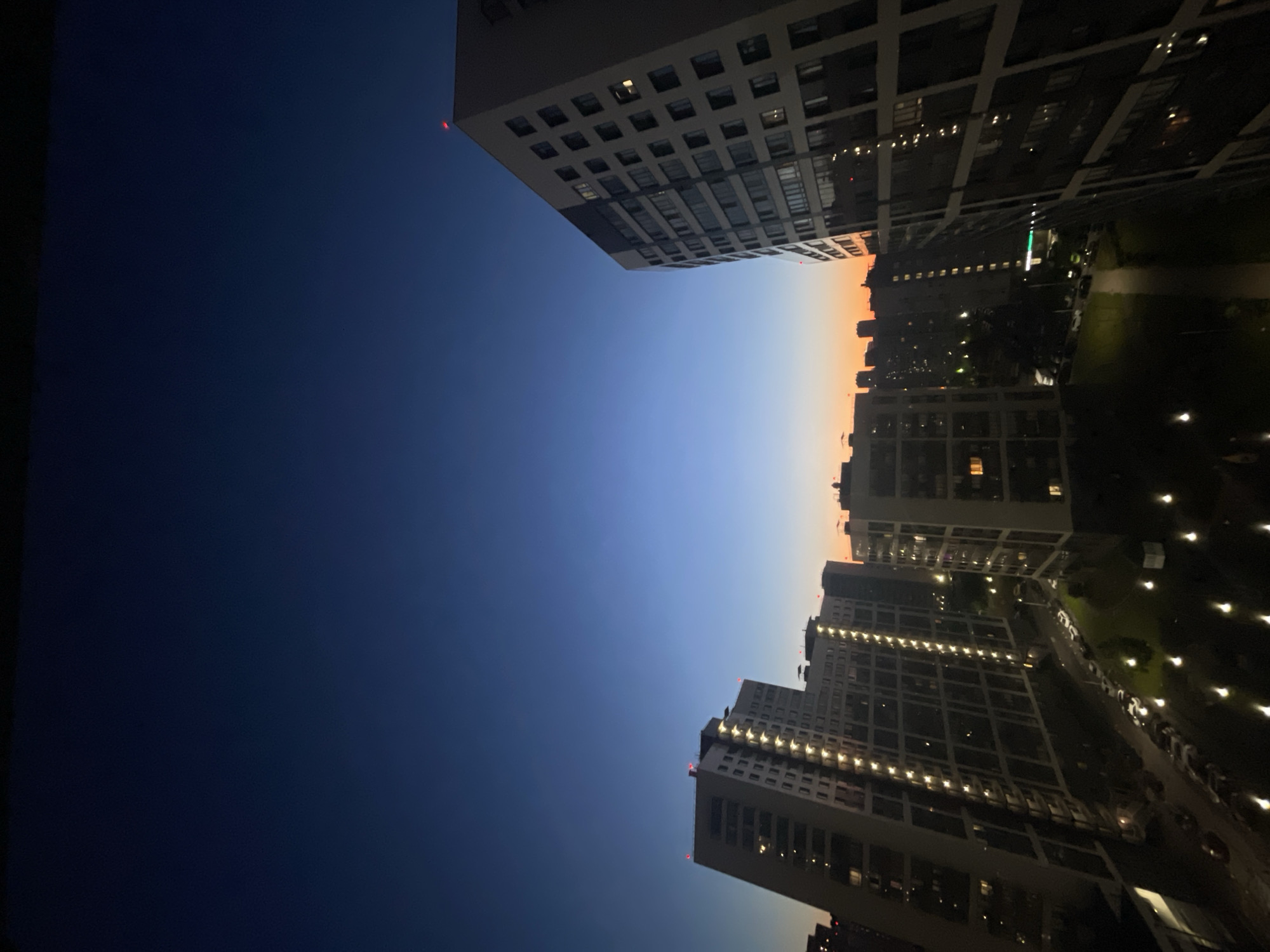The image captures an urban landscape at dusk or dawn. In the foreground, several modern residential buildings with many windows are visible. The buildings are illuminated from within, suggesting occupancy. Some buildings have red lights on their rooftops, likely aviation warning lights. The sky transitions from a deep blue at the top to lighter shades of blue and orange near the horizon, indicative of twilight. A street with parked cars and lampposts is visible between the buildings. The overall scene depicts a residential area in the city of Sinyavino, Russia, during the transitional light of day.