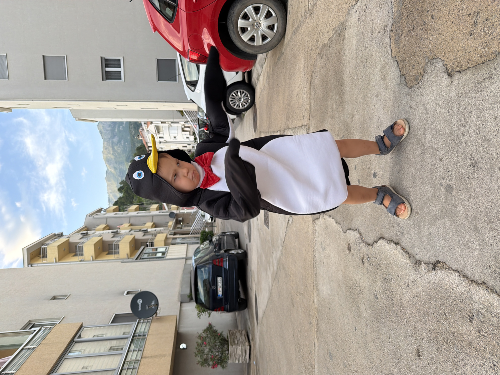 A child in a penguin costume stands on a concrete sidewalk, facing the camera. The child wears blue sandals. Behind the child are buildings, cars, and a mountain range in the distance. The location is Budva, Montenegro.