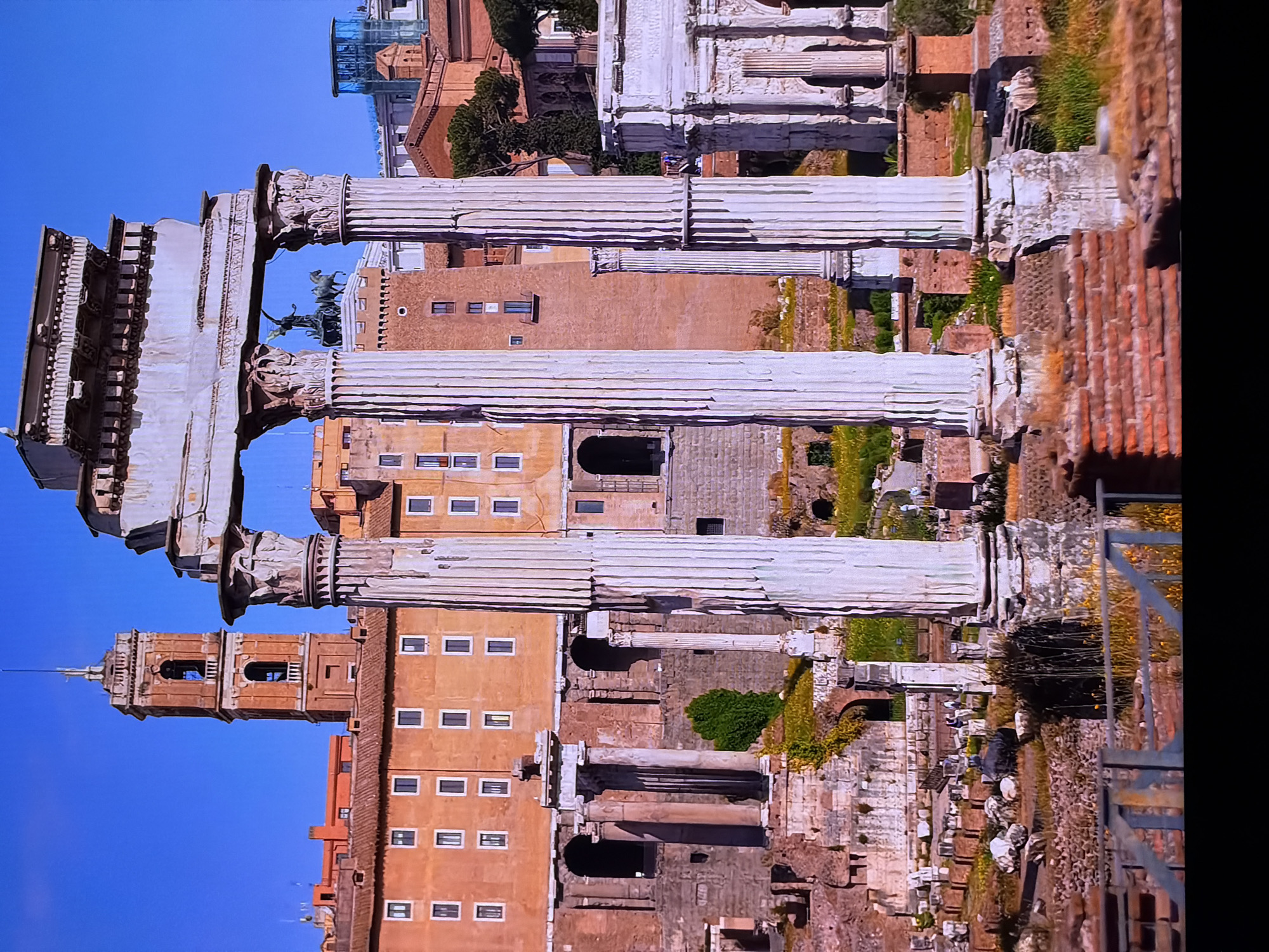 The image depicts the Roman Forum in Rome, Italy, under a clear blue sky. In the foreground, large, weathered marble columns with intricate Corinthian capitals stand tall. Behind them, ancient ruins of stone walls and structures are visible, with patches of green grass and yellow wildflowers growing amongst the rubble.
Further in the background, an orange building with many rectangular windows rises behind the ruins. To the left, a tall bell tower with arched openings stands against the sky. A triumphal arch, also made of stone and partially covered in vegetation, is visible to the right. In the middle ground, a statue of a chariot pulled by two horses can be seen atop a building. The overall scene conveys a sense of historical grandeur and decay. There are no people clearly visible in the foreground.