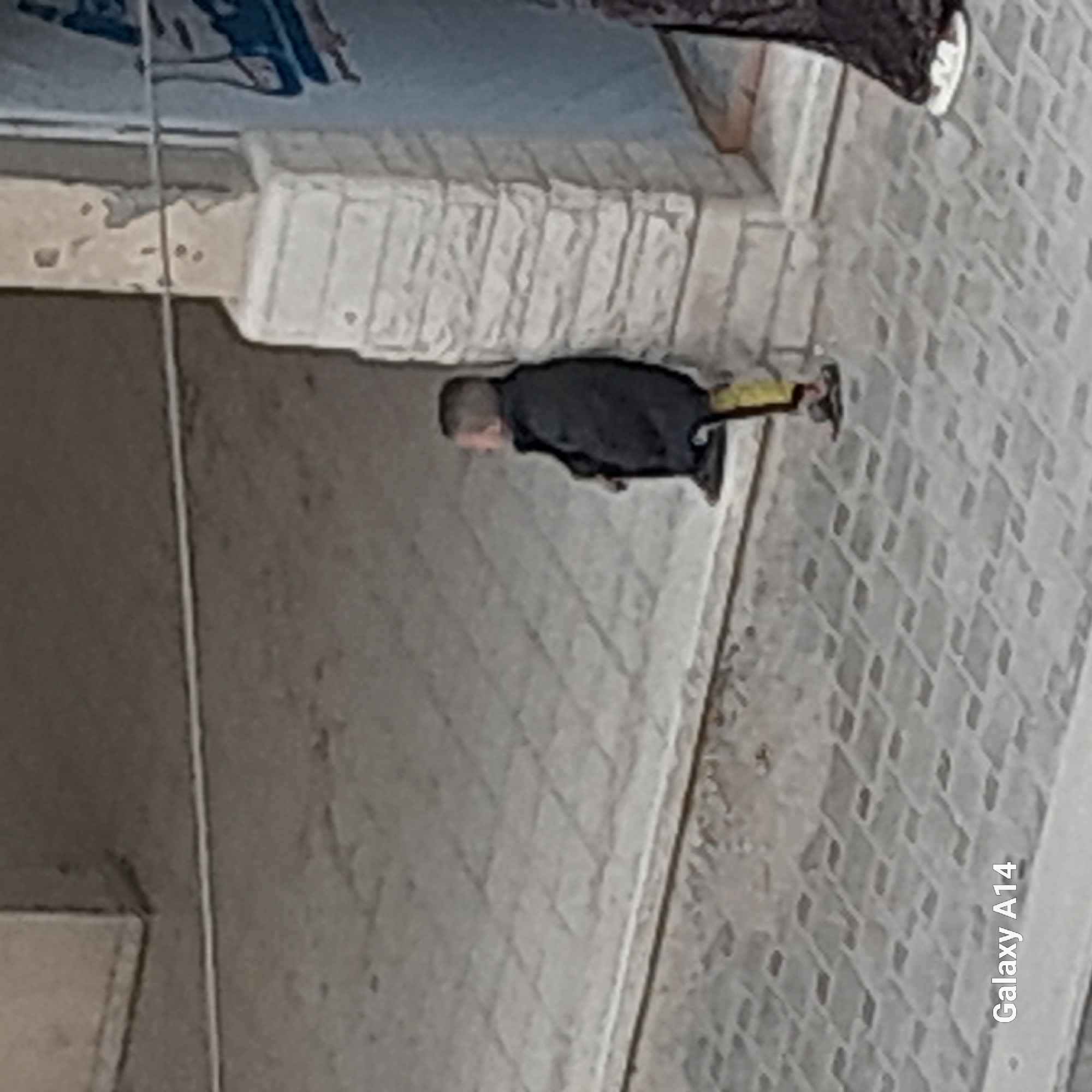 A man is walking near a building in an urban environment. The building has a weathered, textured facade. The man is wearing dark clothing with a hint of yellow. The ground is paved. The scene is possibly taken at a low angle, and the image has a watermark "Galaxy A14". The location and the city are unspecified.