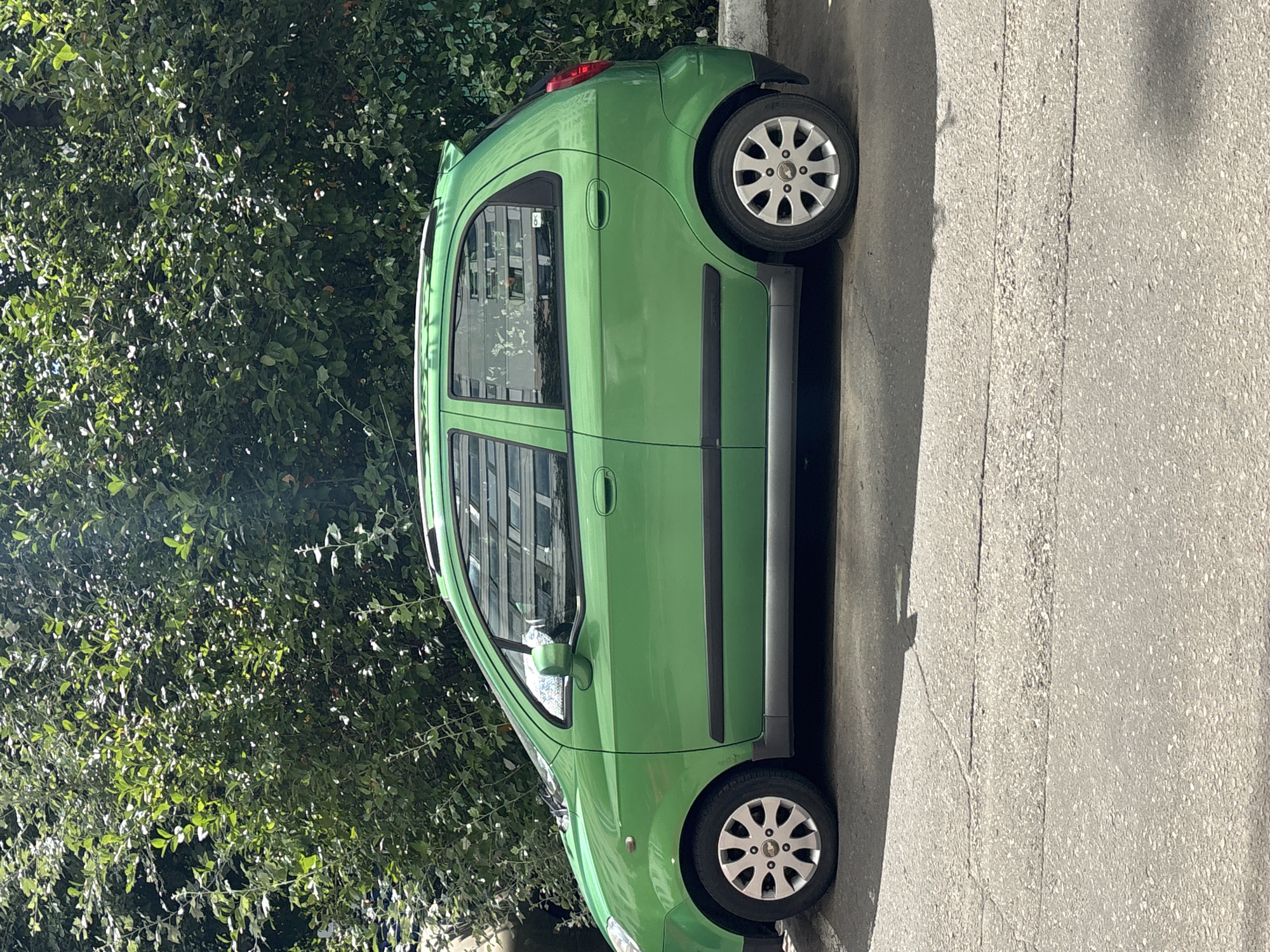 A bright green hatchback car is parked on an asphalt surface next to a dense line of green trees. The car is viewed from its left side. The asphalt shows signs of wear with cracks and patches. The trees appear to be deciduous, with numerous leaves. In the reflection of the car windows, a building with horizontal blinds is visible. The scene appears to be an outdoor urban environment, possibly a street or parking area in Odintsovo, Russia. No people are visible in the image.