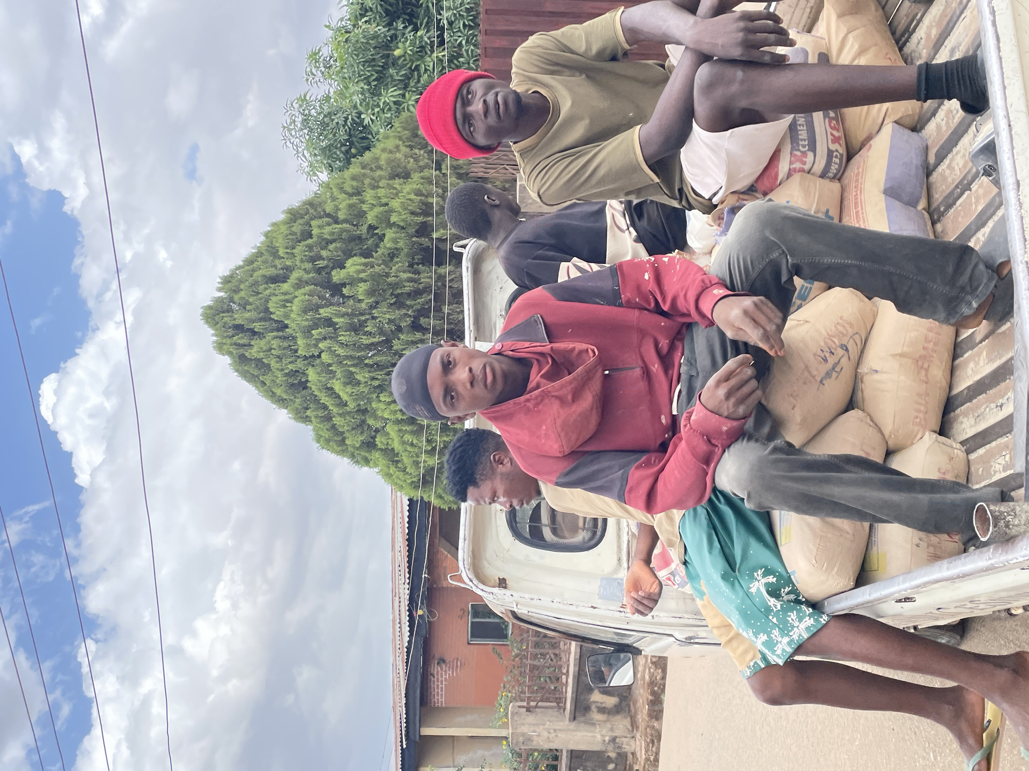 Four young men are seated on bags of cement in the back of a light-colored truck. The truck is parked in front of a building with a corrugated metal roof in Jos, Nigeria. The sky is cloudy. Powerlines are visible above the buildings.