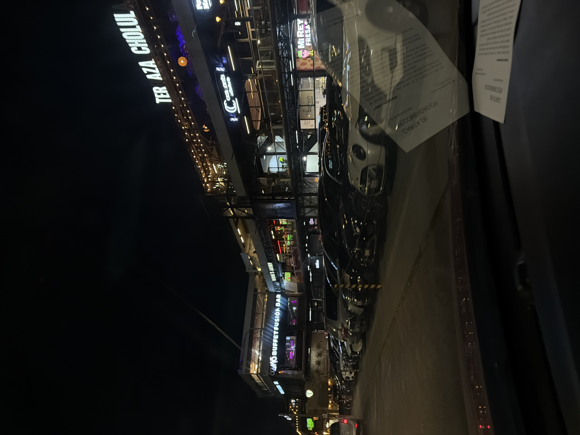 The image shows a nighttime view of a commercial area in San Andrés Cholula, Mexico. The street is lined with several businesses, including restaurants such as "Ter Aza Cholul," "Ozmo Buffet Fusion Bar," "Casa Cortes," and "Mi Rey Fresas." Many cars are parked along the street. The perspective appears to be from inside a vehicle, with some obscured sections due to the windshield.