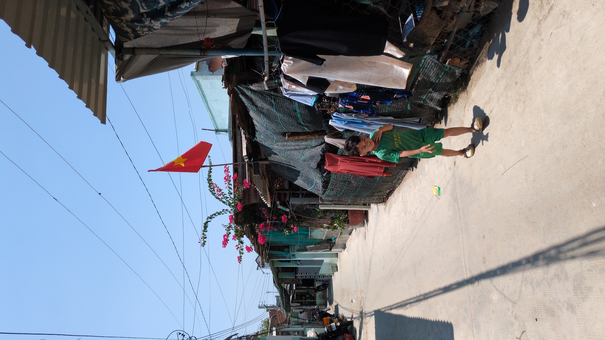 A street scene in Phan Rang – Tháp Chàm, Vietnam. The photo is taken in a narrow street. A child in green clothing stands on a concrete street. Behind him is a residence with clothes hanging on a makeshift line. A Vietnamese flag is visible in the background. Overhead are power lines against a clear blue sky.