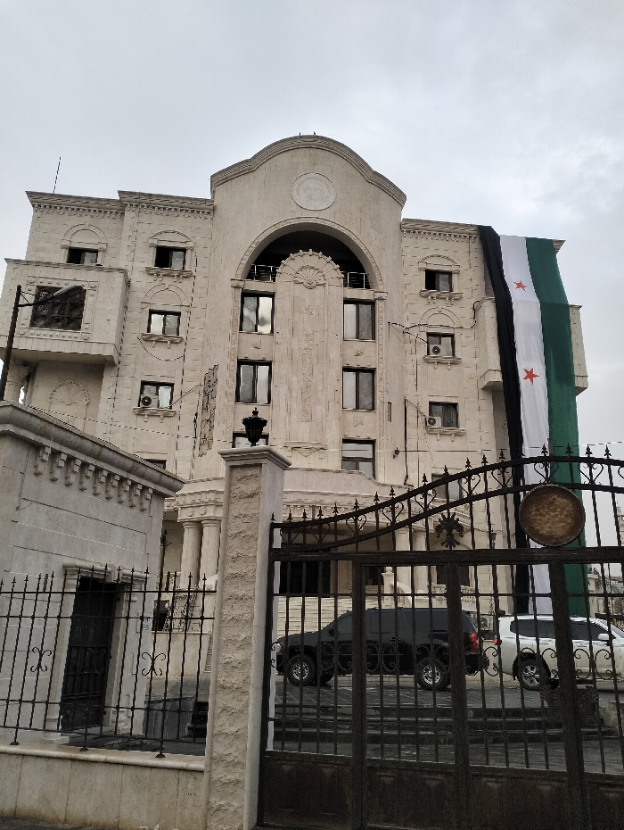 A light stone building with ornate details and arched windows is partially draped with a large Syrian flag. Wrought iron gates stand in front, partially obscuring two parked SUVs, one black and one silver. A smaller stone structure sits to the left, also behind a wrought iron fence. The sky is overcast. The building's architecture suggests an embassy or official government building.