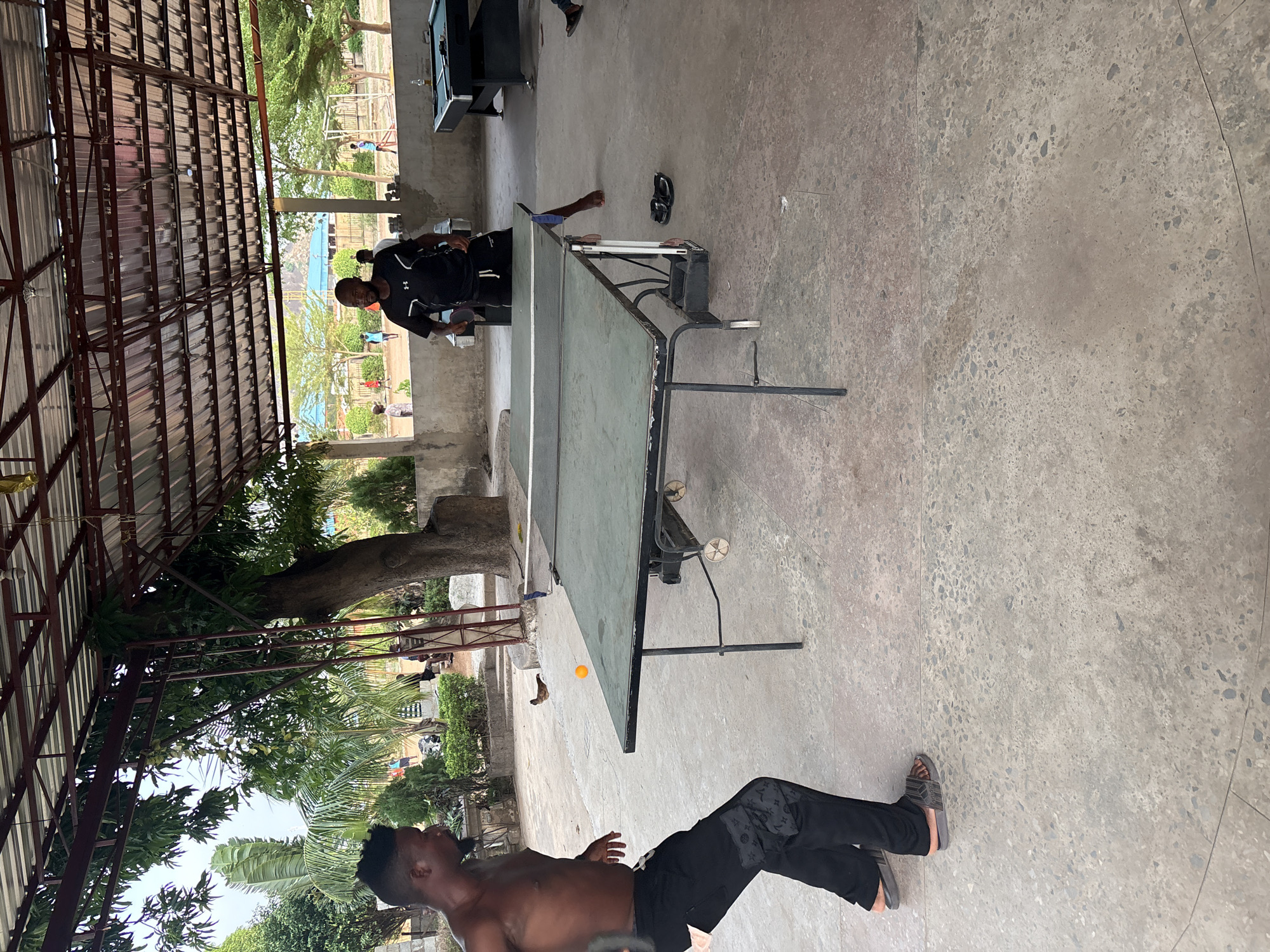The image captures an outdoor recreational scene in Iddah, Nigeria, under a large, open-sided shelter with a corrugated metal roof supported by metal beams. The main activity centers around a game of table tennis (ping-pong).
In the foreground, a shirtless man wearing black pants and gray sandals stands to the left of the ping-pong table. He is looking towards an orange ping-pong ball suspended in mid-air near the ground, suggesting he is actively engaged in play or reacting to a shot.
Opposite him, on the other side of the green ping-pong table, stands another man dressed in a black long-sleeved shirt with an "Under Armour" logo visible on the front, and black shorts. He holds a ping-pong paddle, poised for action. A pair of black flip-flops rests on the light gray, aggregate-textured concrete floor near his position.
Beyond the sheltered area, lush green trees and abundant foliage are visible, indicating a vibrant natural environment. In the background, partially obscured, another dark-colored table, likely a pool table, can be seen, hinting at a multi-functional recreational space. The scene is brightly lit by natural daylight, suggesting it is daytime with clear weather. The overall atmosphere is casual and active.