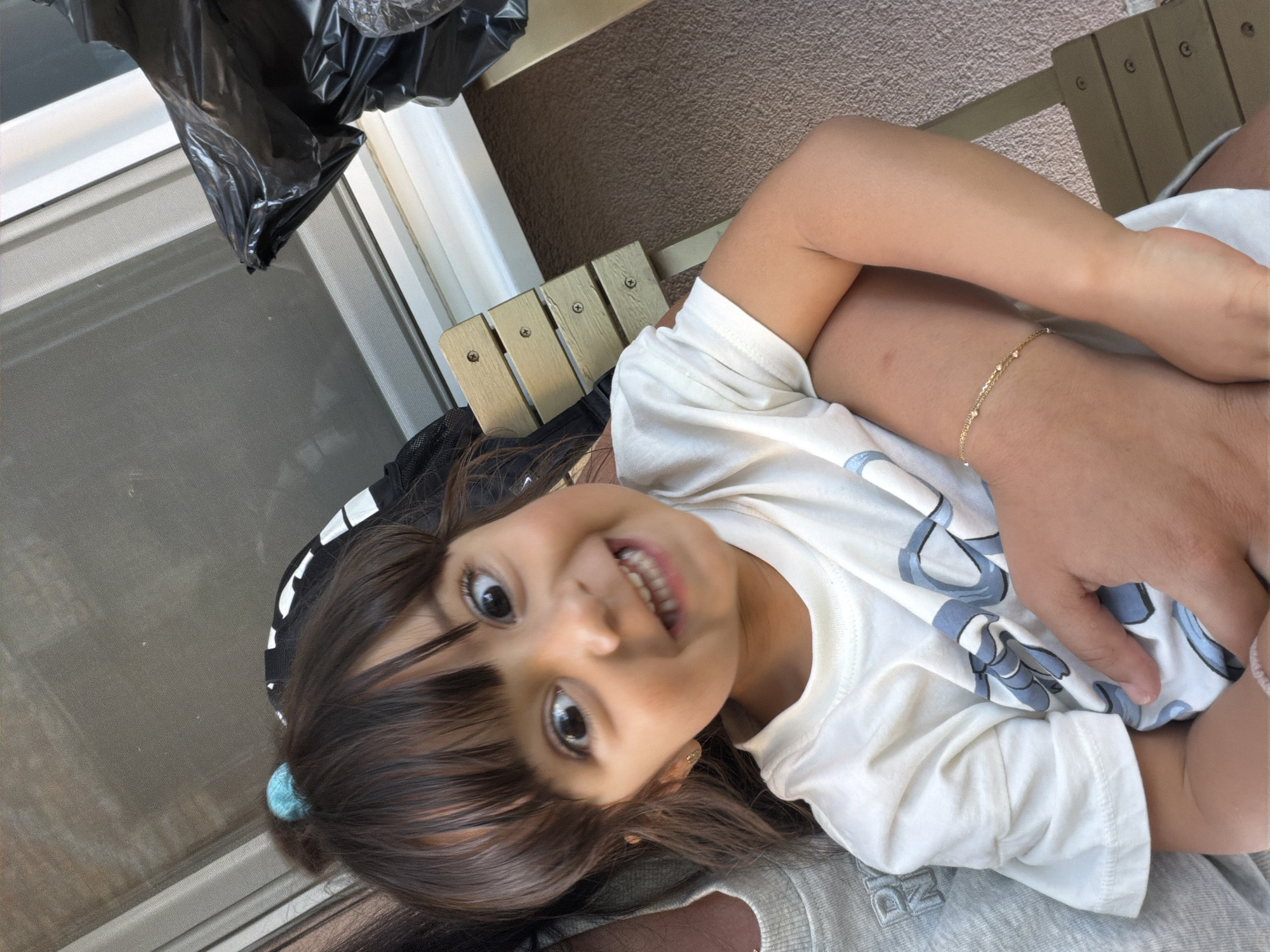 A young girl with dark hair tied up with a blue scrunchie smiles widely at the camera. She is wearing a white t-shirt with a blue graphic print and a delicate gold bracelet on her left arm. The girl is being held by an unseen person, whose arm is visible cradling her. In the background, a wooden bench with three slatted seats is visible. To the right of the bench is a black plastic bag. The overall environment appears to be outdoors, with a window or glass door and possibly a patio area. The image was taken on a sunny day, casting soft light on the scene. The exact location in Burgas, Bulgaria cannot be determined from the visual information present.