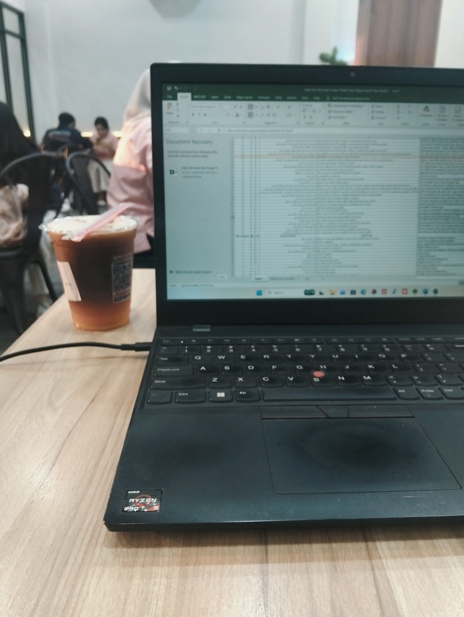 In a bright, indoor environment, likely a cafe in the City of Medan, Indonesia, a black laptop is positioned on a light-colored wooden table. The laptop screen displays an Excel spreadsheet with rows and columns of text, and a "Document Recovery" prompt is visible. To the left of the laptop sits a clear plastic cup filled with a layered iced beverage, possibly coffee with cream, and a pink straw. The background is softly blurred, showing several people seated at tables, some wearing casual attire and one person wearing a light-colored hijab. The overall scene suggests a person working or studying in a public place, accompanied by a drink.
