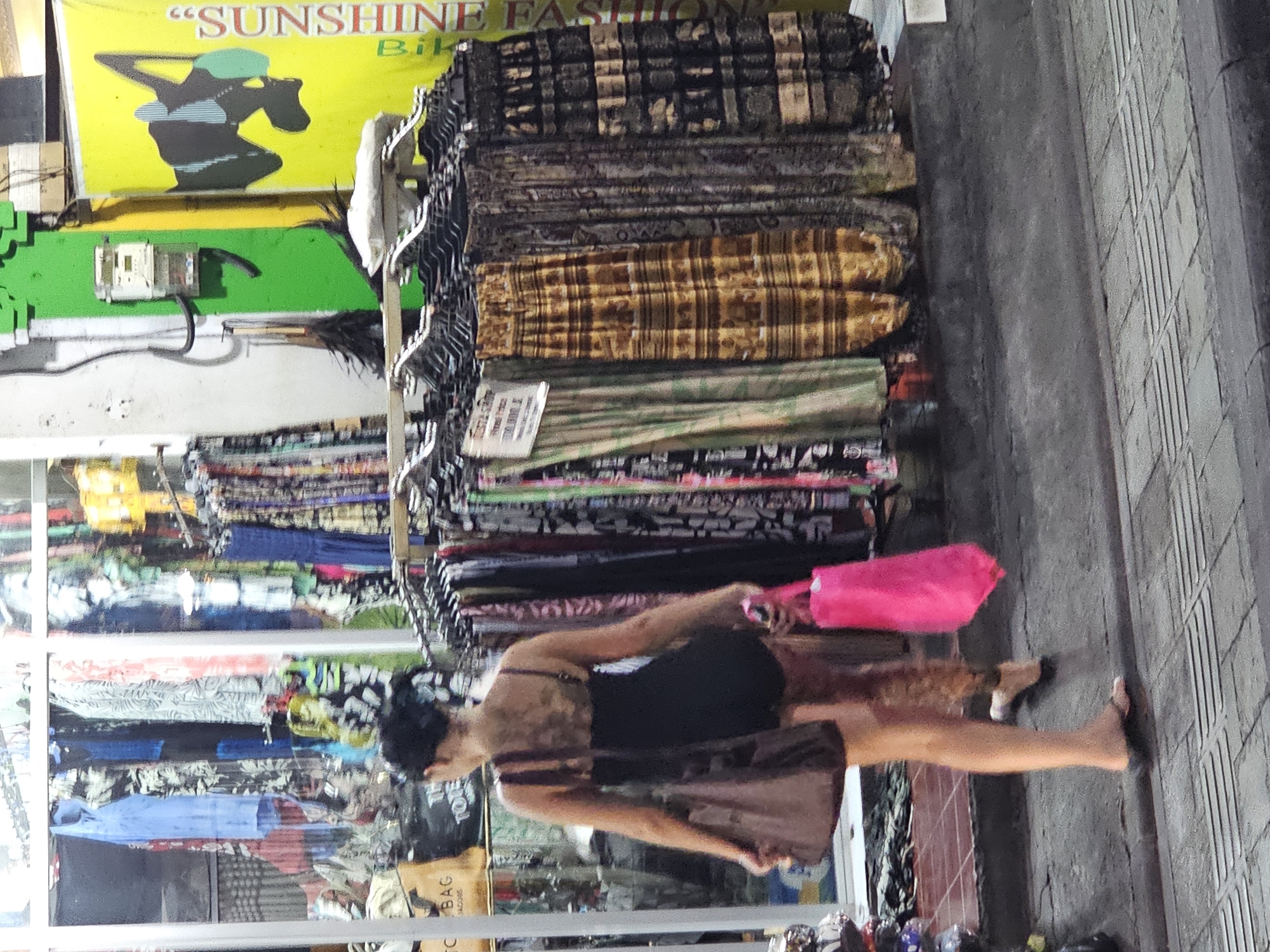 A slightly blurry, medium shot captures a woman in motion, carrying a hot pink tote bag and a brown shopping bag, outside a storefront labeled "SUNSHINE FASHION" in Kuta, Indonesia. The woman is wearing a black dress and is barefoot. Hanging outside the shop is a display of colorful textiles on hangers. The storefront is a glass window with various clothes visible inside. The sidewalk is made of gray brick.