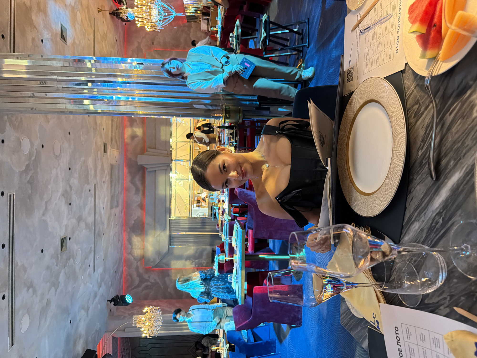 In a brightly lit restaurant in Choeng Thale, Thailand, a woman with dark hair and a black dress sits at a table, looking towards the camera. The table is set with a white plate with a gold trim, wine glasses, a menu, and a plate of watermelon and melon slices. Other people are seated at tables in the background. The restaurant features ornate lighting, a grey-and-white cloud-patterned wall, and mirrored pillars.