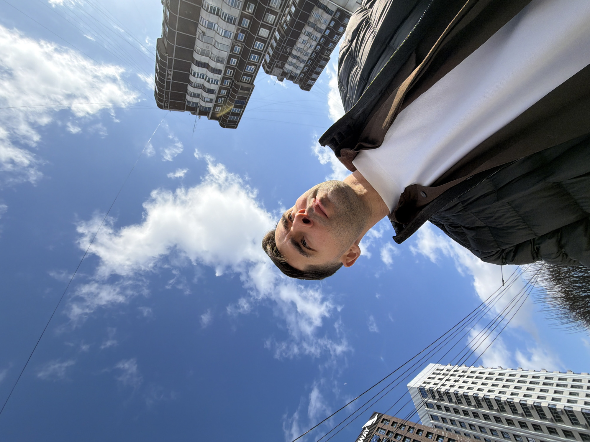 The image is an upward-facing selfie of a man, taken in an urban setting during the daytime. The man's face, visible in the upper central part of the frame, shows him looking directly at the camera with a neutral expression. He is wearing a dark, possibly black, quilted jacket over a white t-shirt.
The background features a bright blue sky with scattered white clouds. Several tall residential buildings frame the sky. A dark-colored apartment building with numerous windows is prominent in the upper left, while a lighter, modern high-rise building (white/grey) with many rectangular windows is visible in the lower right. Adjacent to this white building, a darker brown building is partially seen. The architecture suggests a contemporary urban environment, consistent with Krasnoyarsk, Russia.
Multiple thin, dark wires crisscross the sky, indicating utility lines common in cities. A small, bare tree or bush top is visible on the far right edge of the frame. The lighting suggests it is a sunny day. On the white building in the bottom right, the text "CITYWAY" is discernible on a dark sign. The overall perspective is from a low angle, emphasizing the height of the buildings and the expansive sky.