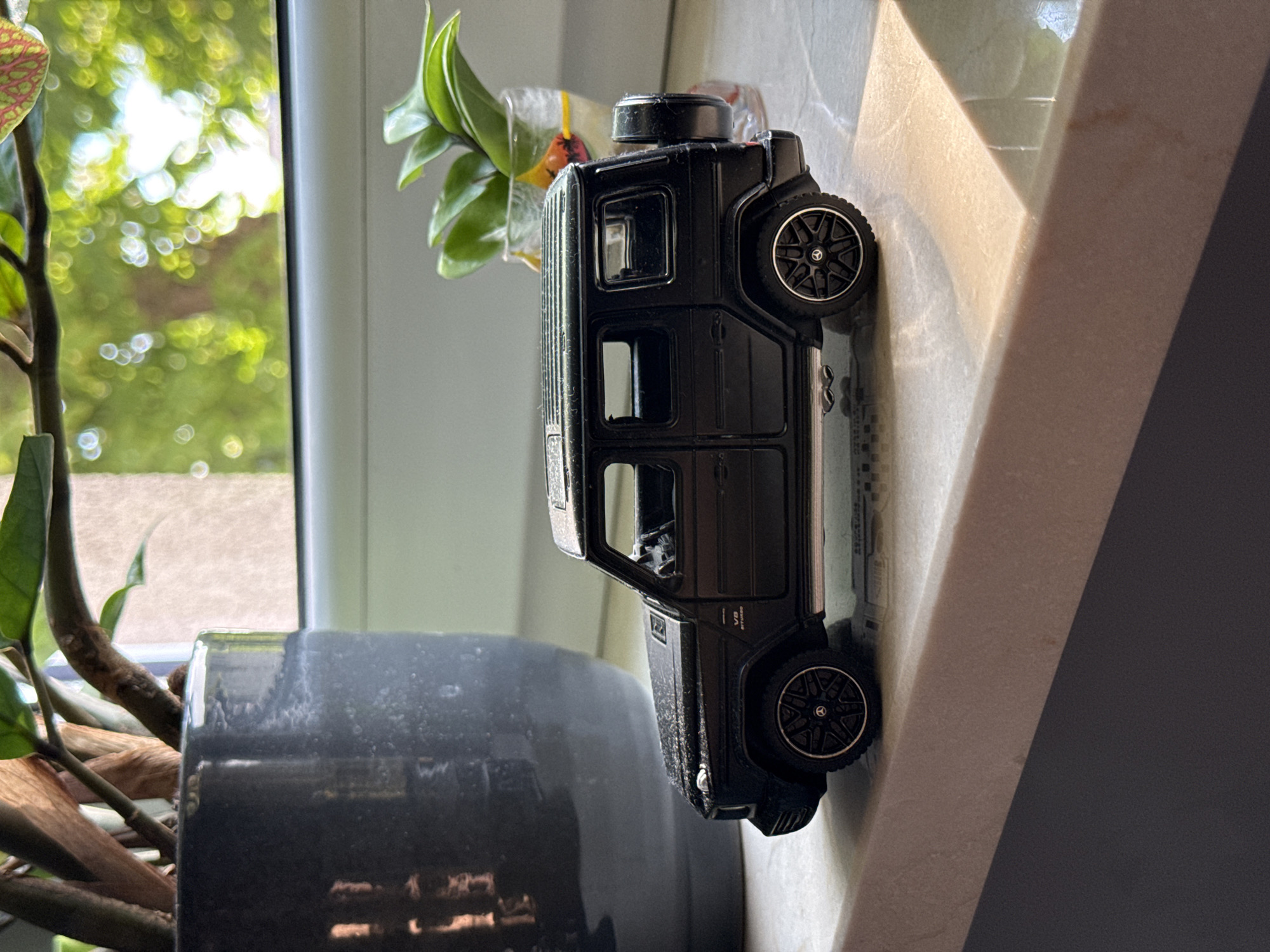 A black toy Mercedes-Benz G-Wagon is positioned on a marble ledge. Behind the toy car, a large dark grey planter with a plant is visible. A glass container with a plant is to the right of the toy car. The background shows a window with dappled sunlight filtering through green foliage. The scene appears to be indoors, possibly on a windowsill. The location in the city of Łódź, Poland is not discernible from the image.