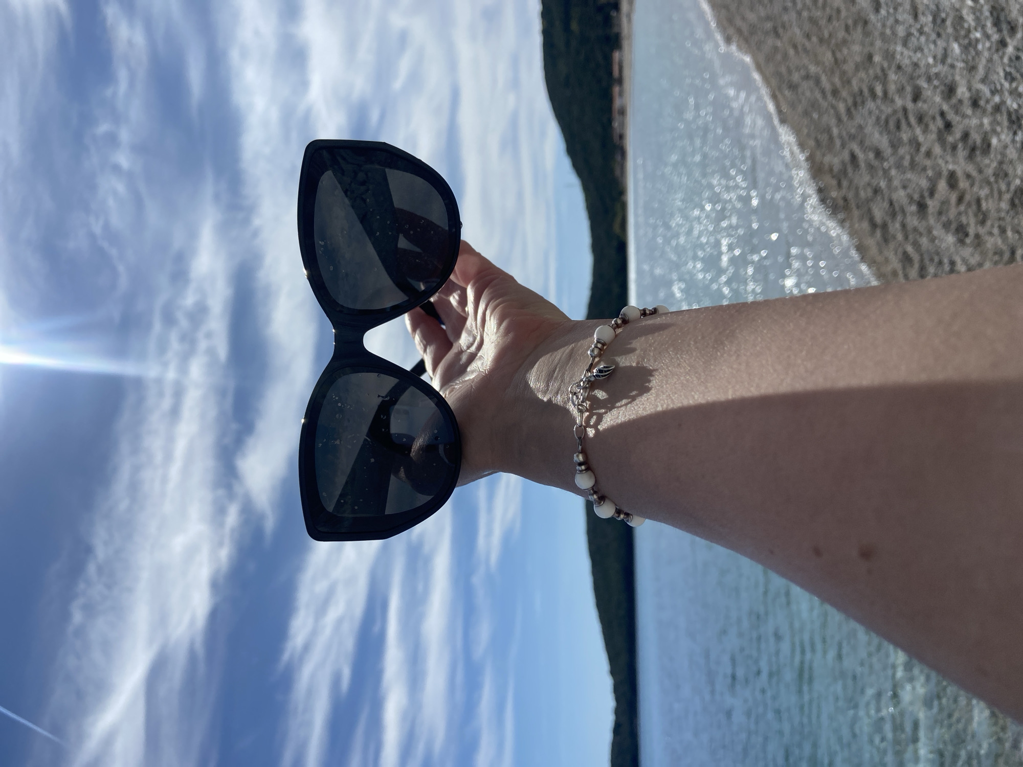 A person holds sunglasses over a view of the Bay of Kotor in Prijevor, Montenegro. The sunglasses are dark-framed, with visible smudges on the lenses. The person's hand and forearm, adorned with a beaded bracelet, are visible. The bay is sunlit, with light reflecting on the water. A forested area is visible across the bay under a sky with scattered clouds.