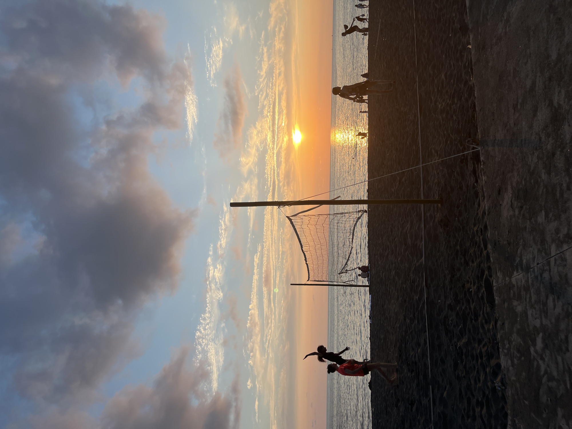 The image depicts a beach volleyball game at sunset on the black sand beach of Ureki, Georgia. The sun is low on the horizon, casting a warm glow across the sky and water. Several people are playing or watching volleyball, with a net set up on the sand. The sea is calm, and the sky is filled with colorful clouds.