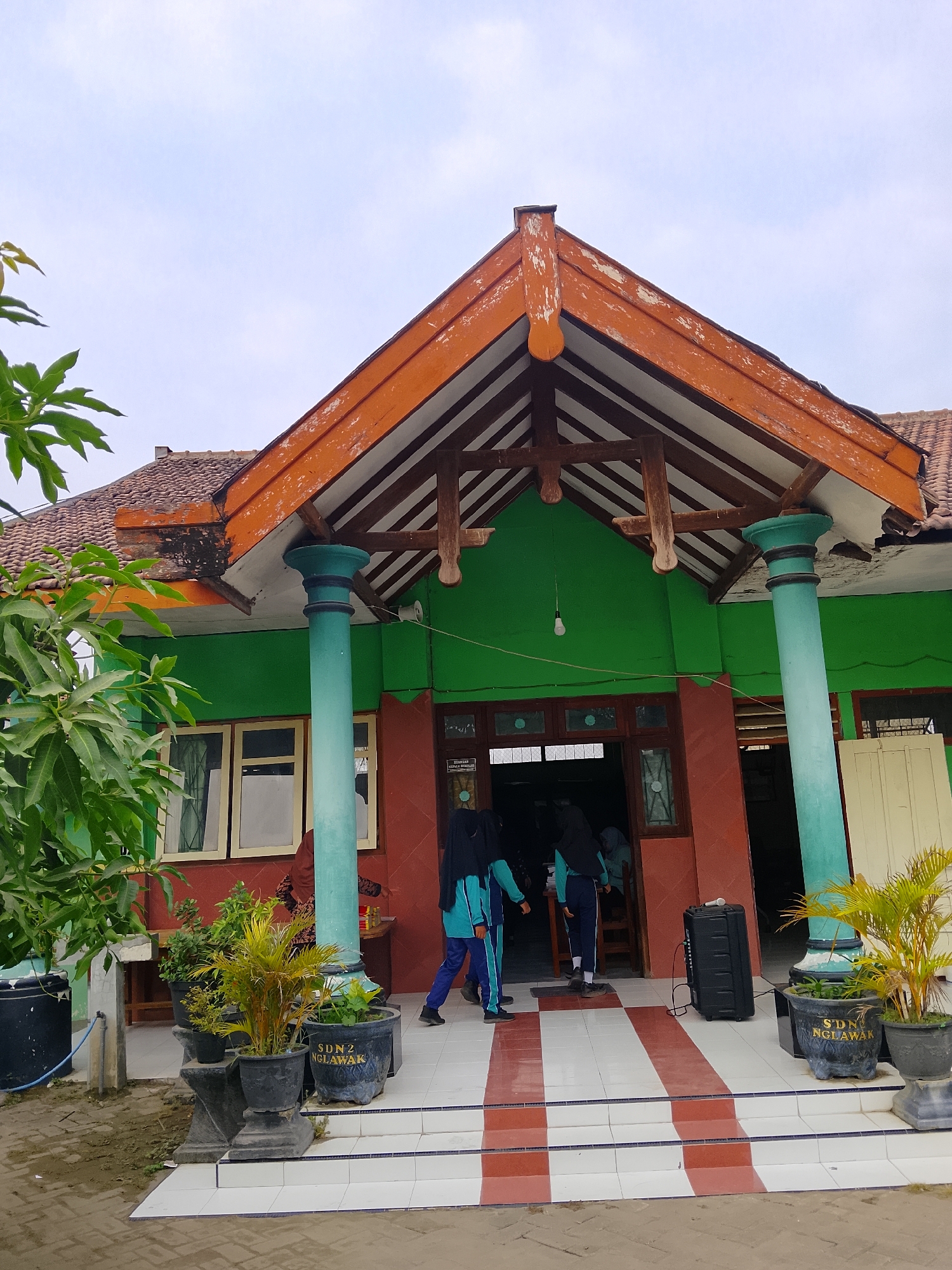 This is a medium shot of the entrance to a school building, likely in Kepuh, Indonesia, given the visible text on the planters. The building features a prominent green facade with a reddish-brown lower section. Above the entrance, an open-air wooden roof structure with orange-painted trim is supported by two large, teal-colored columns. The roof itself has traditional terracotta tiles.
In front of the building, there are several potted plants. One large black planter prominently displays the text "SDN 2 NGLAWAK" in yellow lettering, indicating it is associated with the school. Two individuals, dressed in what appear to be school uniforms including headscarves, are walking towards the entrance. One is wearing a teal and black uniform, and the other is wearing a teal and blue uniform. A large black portable speaker sits on the tiled steps leading up to the entrance.
To the left of the building, a large green leafy tree is visible, partially obscuring the upper portion of the building. The ground in front of the steps is paved with interlocking bricks. The sky is overcast. The overall impression is of a modest, functional school building.