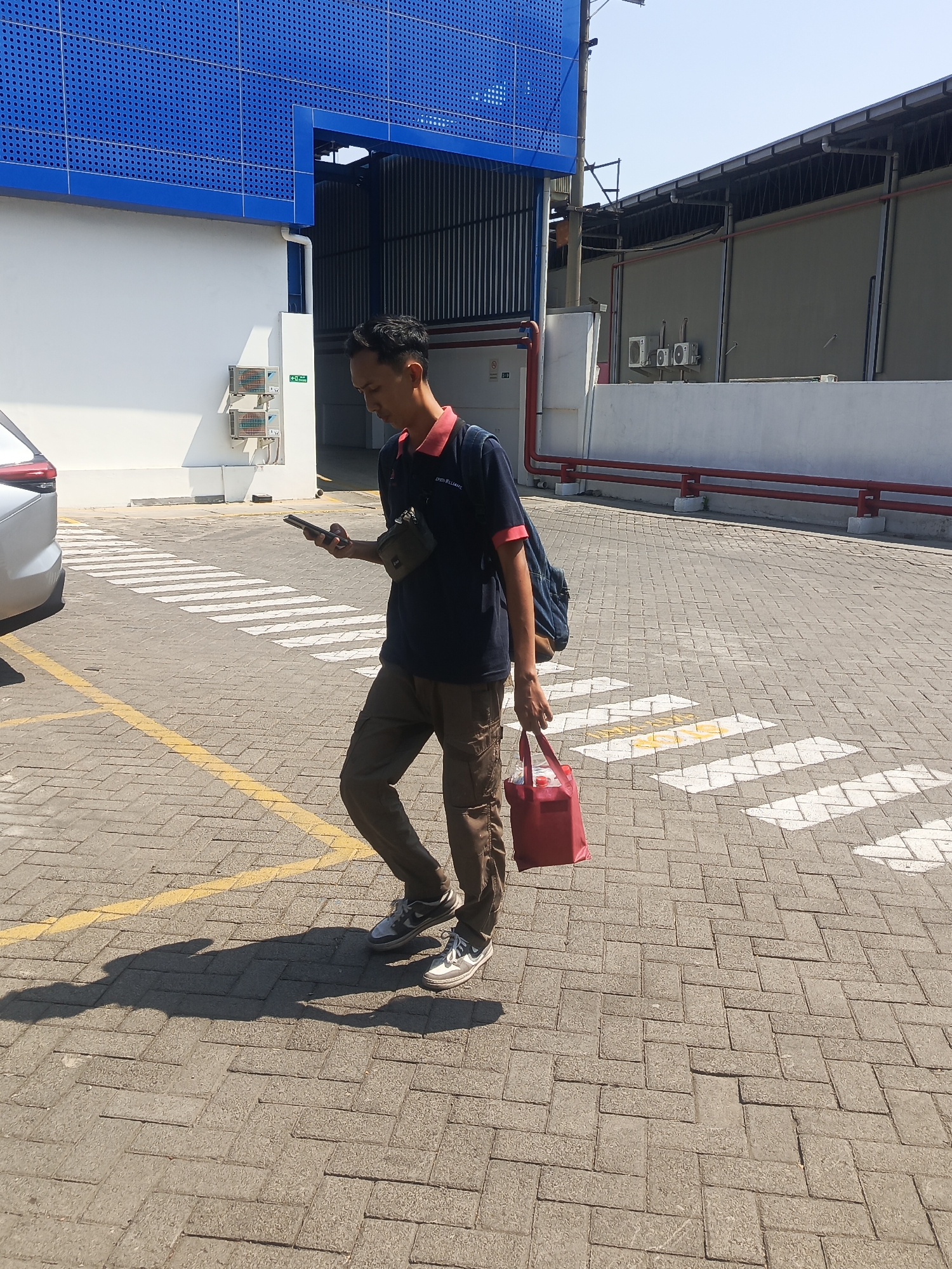 A man is walking on a paved area in Wadungasih, Indonesia, looking at his phone. He is wearing a dark blue polo shirt with a red collar and dark brown pants. He has a small green bag slung across his chest and a red tote bag in his right hand. He is carrying a blue backpack on his left shoulder. The ground is made of grey interlocking pavers, with white and yellow crosswalk markings visible. In the background, there is a white building with a blue facade featuring a pattern of circular holes, and a grey building with red pipes running along its exterior. The sky is clear and blue, indicating a sunny day.