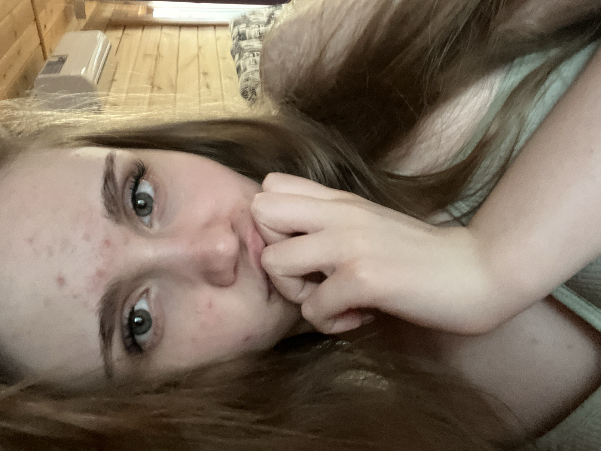 A young woman with long, reddish-brown hair and light-colored eyes is looking at the camera. She has blemishes on her forehead and cheeks. Her left hand is raised to her mouth, with her fingers touching her lips. She is wearing a light-colored garment. The background appears to be a wooden room with an air conditioner mounted on the wall. The image was taken in the city of Ashkadar, Russia.