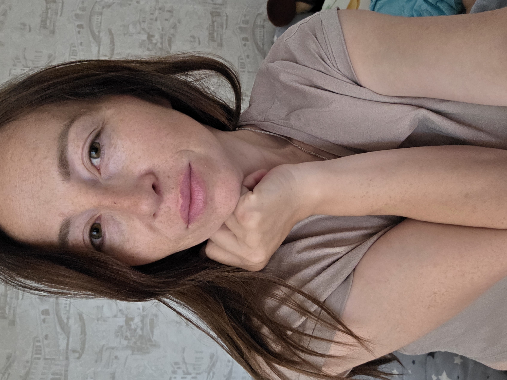 This is a head and shoulders portrait of a woman with brown hair and fair skin, wearing a light brown t-shirt. Her right hand is resting on her chin, and she is looking directly at the camera. The background is a wall with a patterned wallpaper. The image appears to have been taken indoors, possibly in a residential setting. There is no indication of the city of Chaikovsky, Russia.