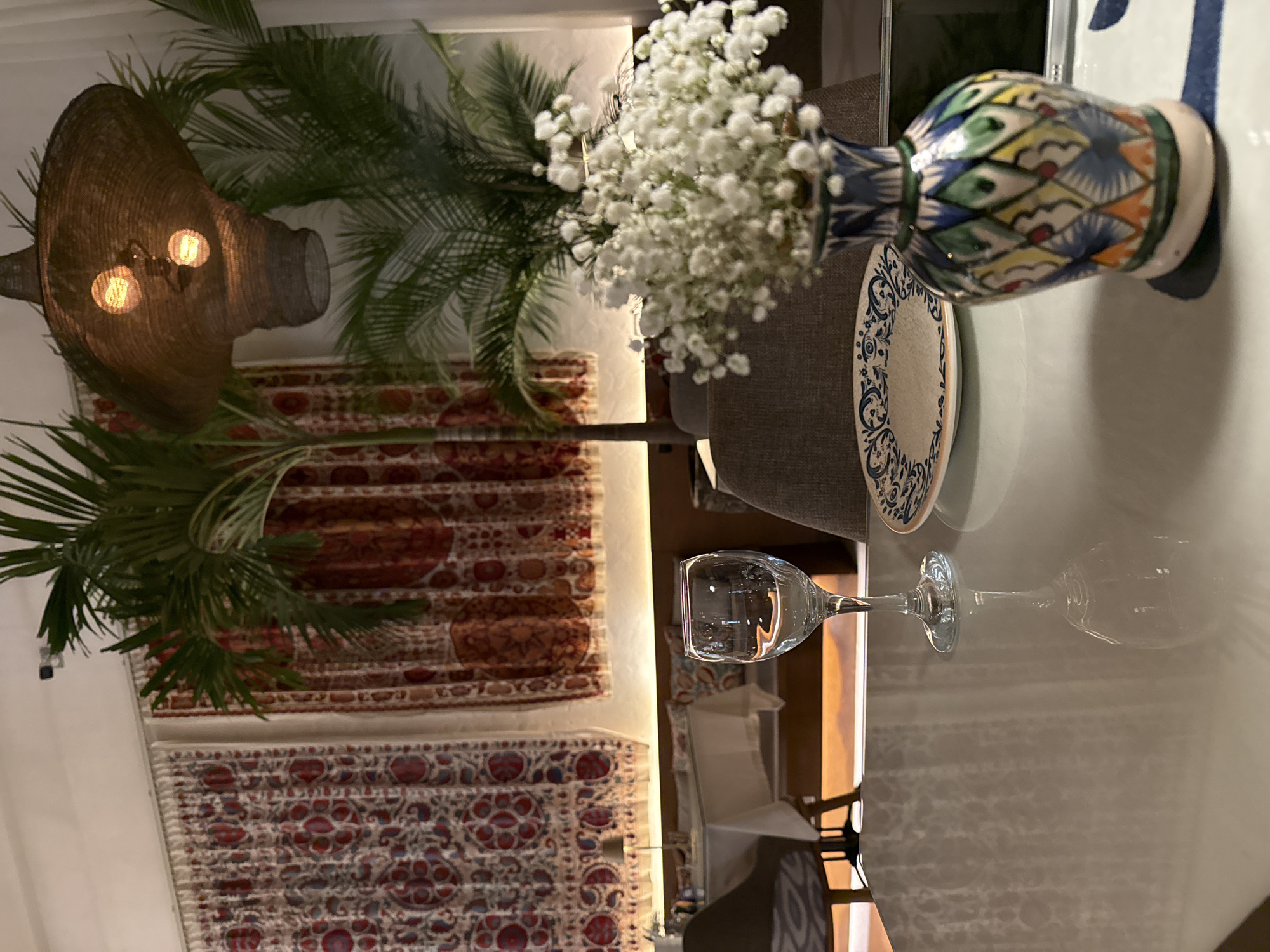 The image shows a table setting in what appears to be an interior space, potentially a restaurant or a dining area in a home. The table is set with a plate, a decorative vase containing white flowers, and a water glass. There is a potted plant with palm-like leaves. In the background, there are decorative tapestries or wall hangings featuring complex patterns and colors. A woven lamp is visible. Based on the style and ambiance, the location might be in Dubai, United Arab Emirates.