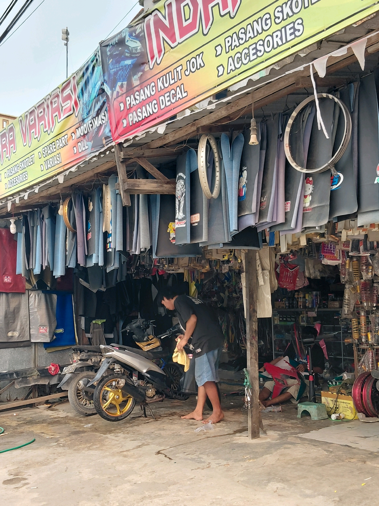 A street-level view of a motorcycle accessory shop in Persiapan Sumber Sari, Indonesia, captures a bustling scene. The shop's facade is adorned with a large, colorful banner advertising "VARIASI" and various services such as "PASANG SKOTLET," "MODIFIKASI," "SPARE PART," "DLL," "PASANG KULIT JOK," "PASANG DECAL," and "ACCESORIES." Hanging from the shop's roof are numerous motorcycle seat covers, some displaying cartoon characters like Mickey Mouse and Hello Kitty, alongside plain black and blue ones. A single light bulb dangles from the roof, illuminating the cluttered interior.
In the foreground, a man in a black t-shirt and blue shorts is actively cleaning a silver motorcycle with a yellow cloth. Another individual is seated or resting on the ground in the background, surrounded by motorcycle parts and supplies. The ground is concrete, showing signs of wear and tear, with a green hose lying on the left side. The overall environment appears to be an outdoor, open-air market or roadside stall, common in Indonesian cities. The scene is filled with an array of motorcycle parts and accessories, including shock absorbers, rims, and various plastic components, all arranged for sale or repair. The lighting suggests it is daytime, with the sky appearing overcast.