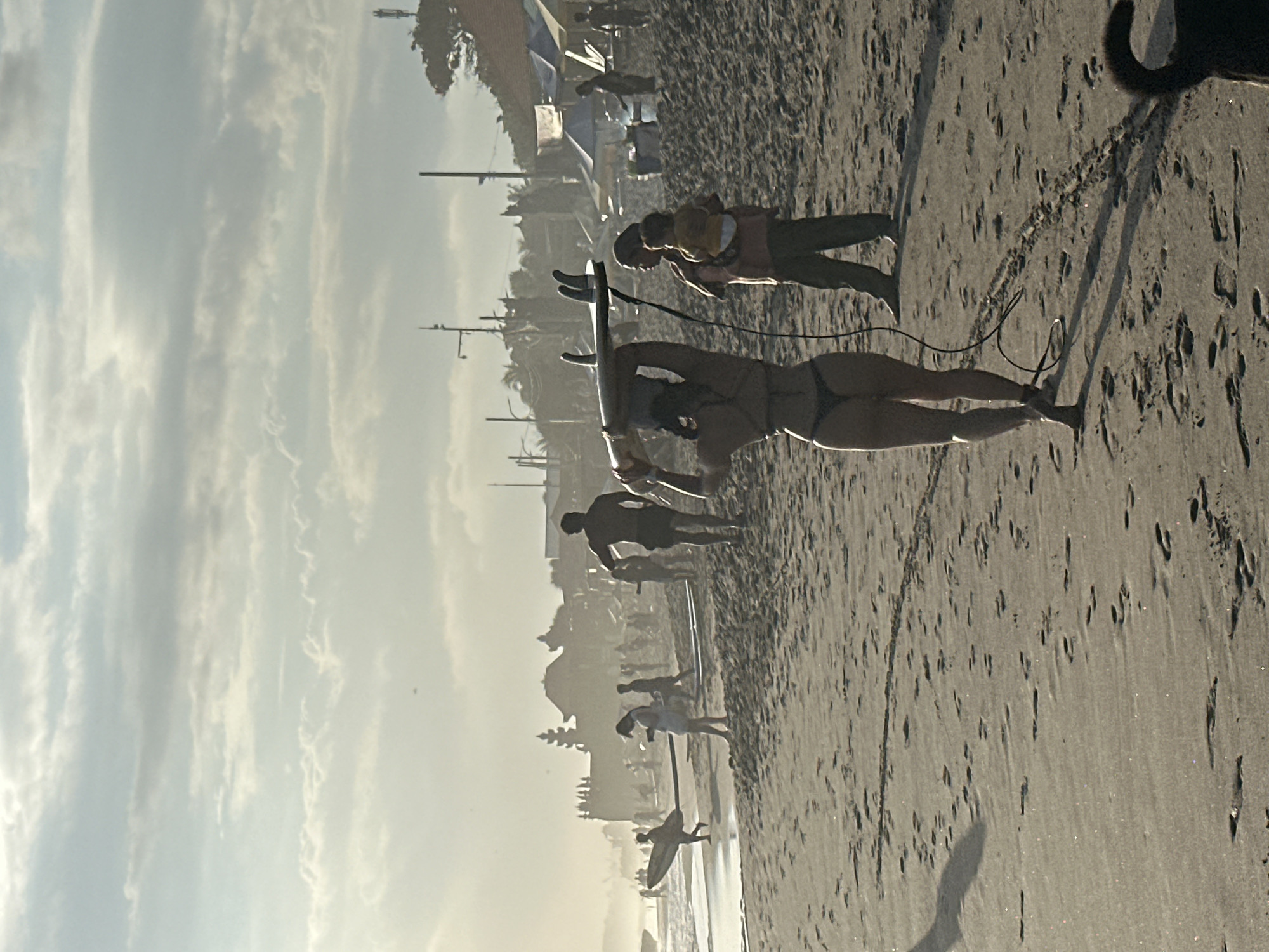 An outdoor, slightly hazy, over-the-shoulder medium shot captures a beach scene in North Kuta, Indonesia, during either sunrise or sunset. The sand is littered with footprints, and a dark shadow of a person, holding a surfboard overhead, falls across the beach from the left. In the foreground, a woman in a bikini carries a surfboard on her head, facing away from the camera. Beside her, a person in dark pants holds a child on their hip. In the midground, other figures are visible on the beach, some with surfboards, and tents and buildings line the distant shore, partially obscured by haze. The sky above is filled with scattered clouds, with a hint of palm trees and utility poles visible on the right side.