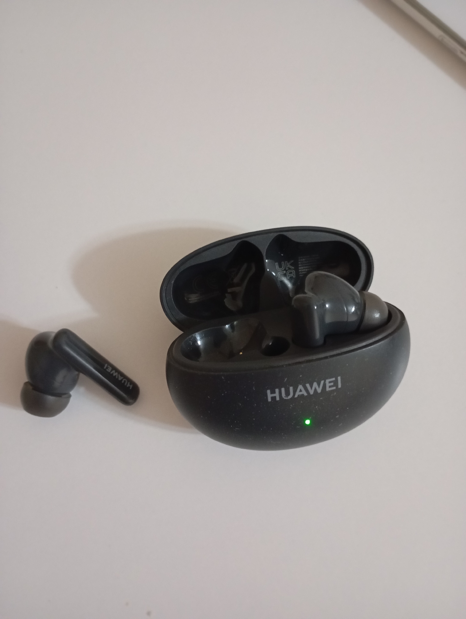 In an indoor, well-lit setting, possibly a desk or table, a pair of black Huawei FreeBuds are depicted. One earbud rests on its side to the left of an open charging case. The charging case is positioned to the right, with its lid open, revealing two earbud slots. The earbud on the left displays the "HUAWEI" logo in white text. Inside the charging case, another earbud is partially visible, along with some faint text indicating "UK CA". A small green LED indicator light is illuminated on the front of the charging case. The background is a plain, light-colored surface. There is no information or visual cues that indicate the location in the city of Goryachiy Klyuch, Russia.
