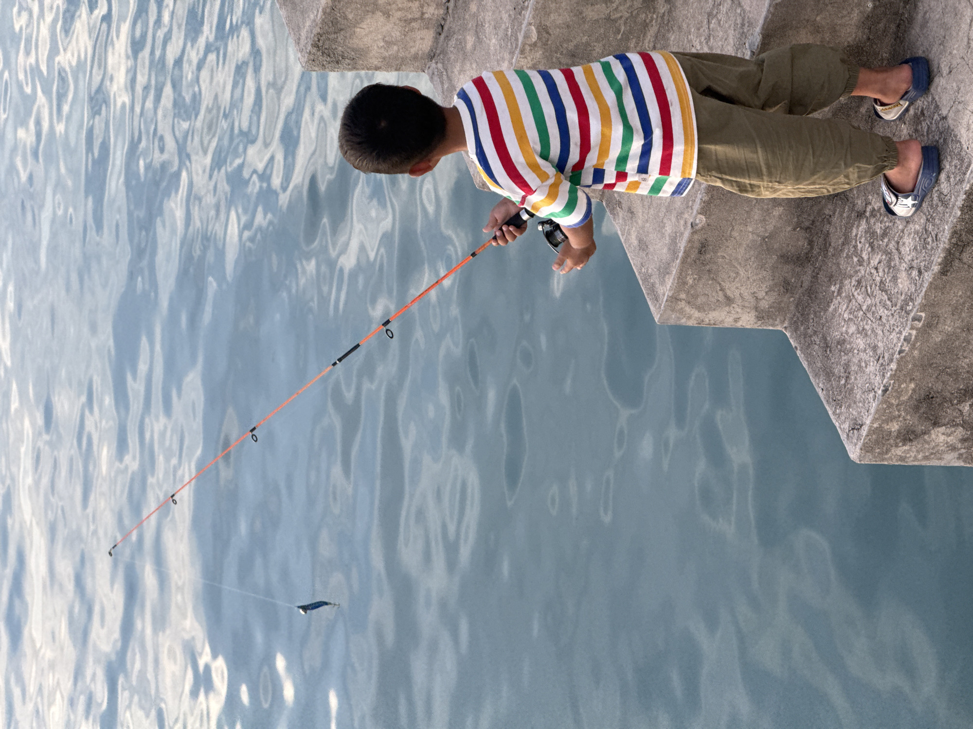 A young boy stands on concrete steps, facing a body of water, holding a fishing rod with his gaze fixed on the water. The boy is wearing a horizontally striped t-shirt in red, yellow, green, and blue, olive green pants, and sandals. The fishing line extends from the rod into the water, where a blue lure is visible. The water exhibits ripples and reflections. The concrete steps appear weathered. The scene is likely outdoors.
