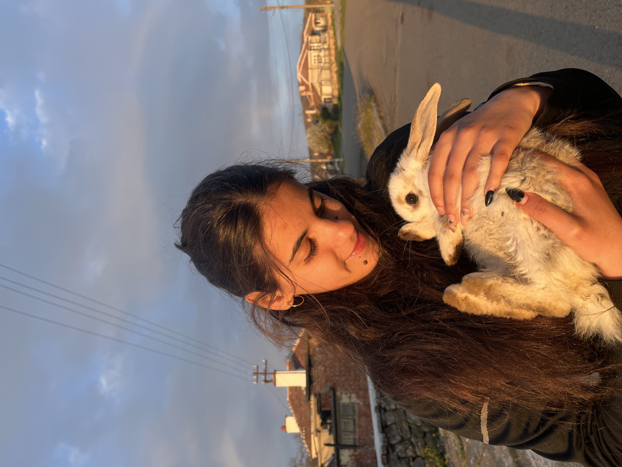 A woman is holding a rabbit outdoors. The woman has dark hair and is wearing a black jacket. The rabbit is light brown and white. The scene appears to be in a residential area with houses in the background. There are utility poles and wires visible against a cloudy sky. Based on the setting, this photograph was taken in the town of Lozarevo, Bulgaria.
