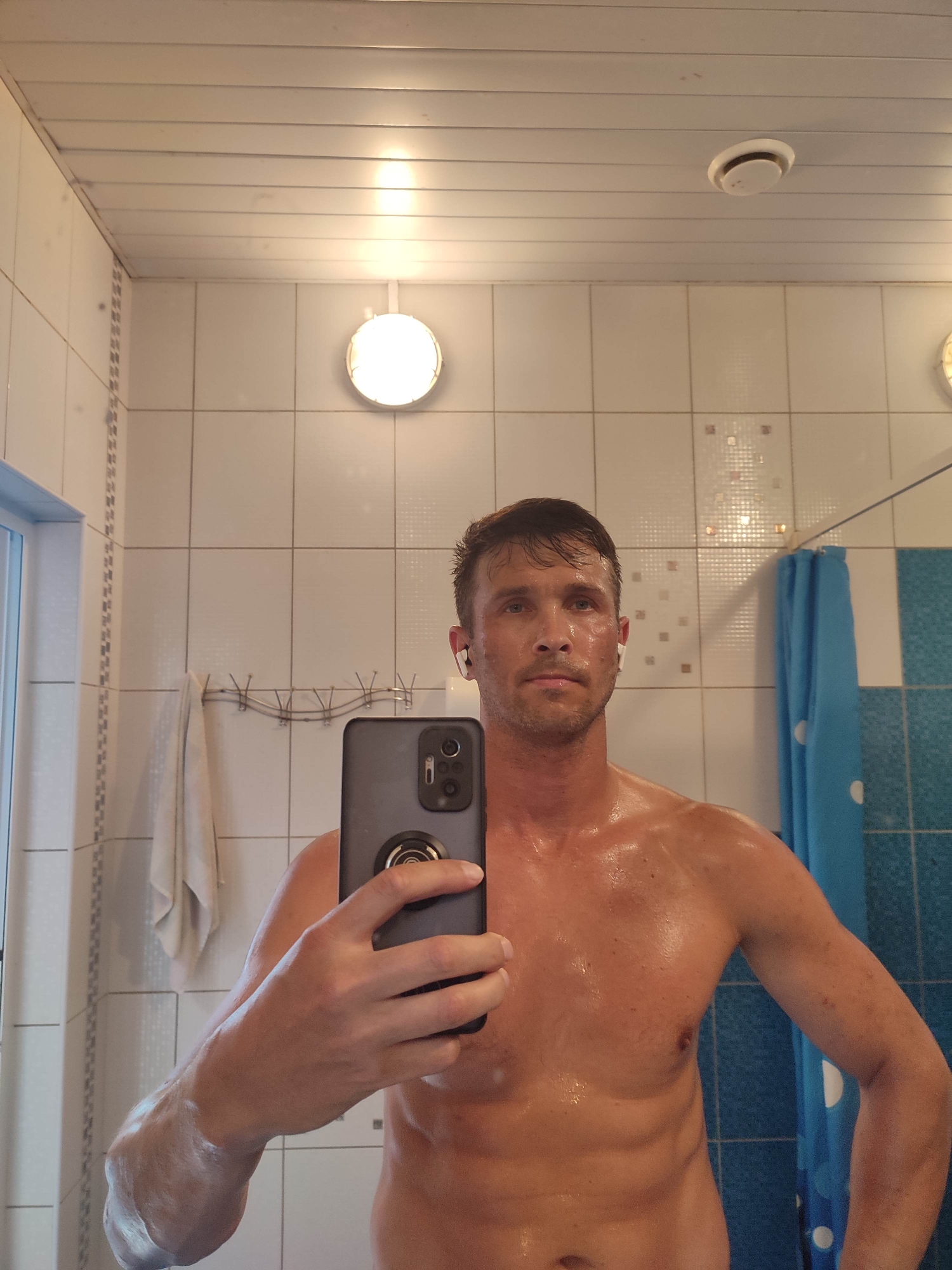 A man is taking a mirror selfie in a bathroom. He is topless and appears to be sweating, with water droplets visible on his skin. He is holding a smartphone in his right hand, which is pointed towards the mirror. He is wearing white earbuds. The bathroom has white tiled walls and a white ceiling. A shower curtain with blue and white polka dots is visible in the background. A towel hangs on a rack on the wall. The scene appears to be a domestic bathroom, not specifically identifiable as being in a particular city or location.