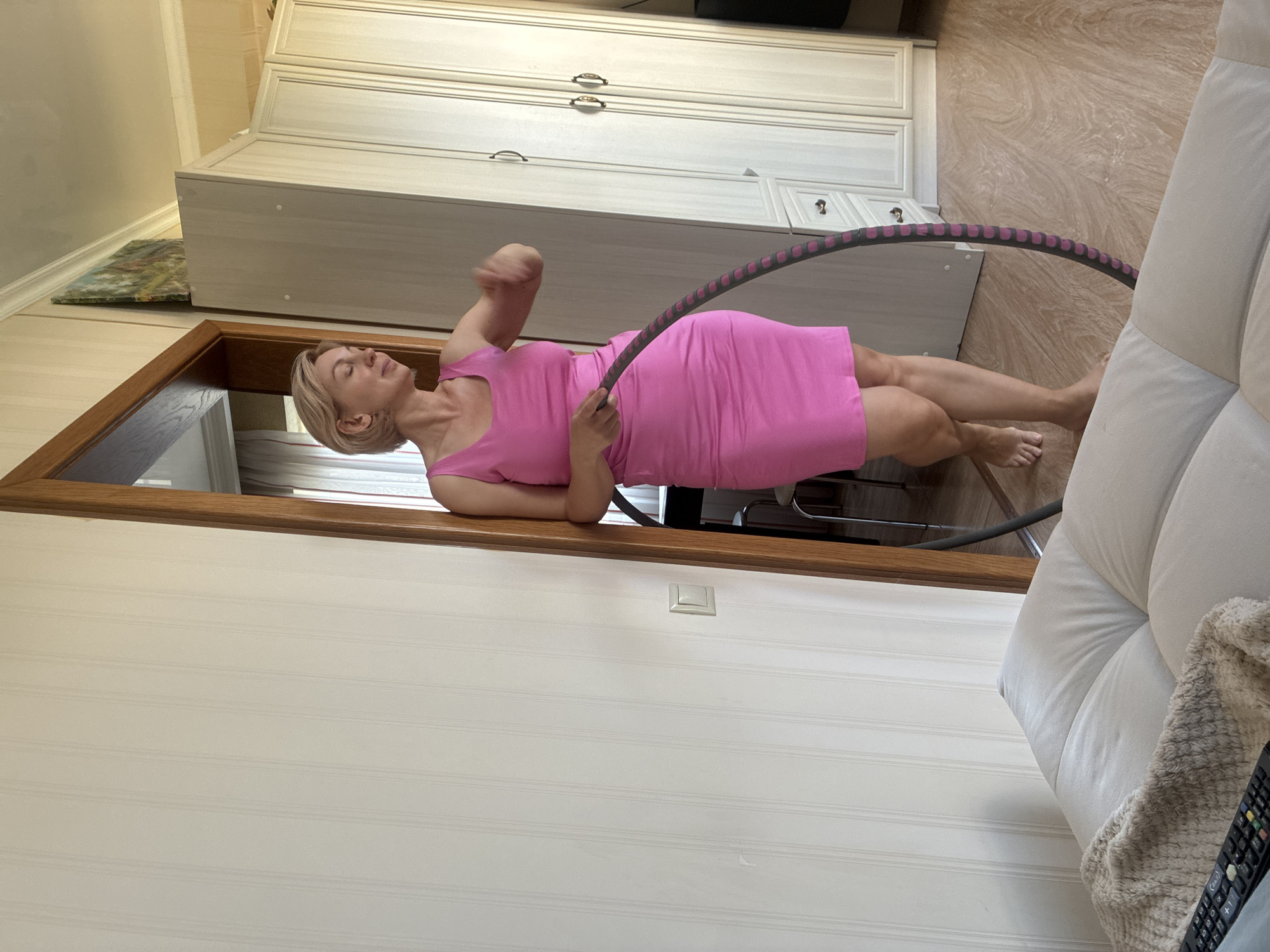 A woman with blonde hair, wearing a pink dress, is lying on her back on the floor, inside a doorway. She is holding a hula hoop above her. The room has light-colored walls and wooden trim. In the background, there are white cabinets with dark handles. To the right of the woman, a corner of a white couch is visible. The floor appears to be wood. The scene suggests an indoor activity, possibly exercise.