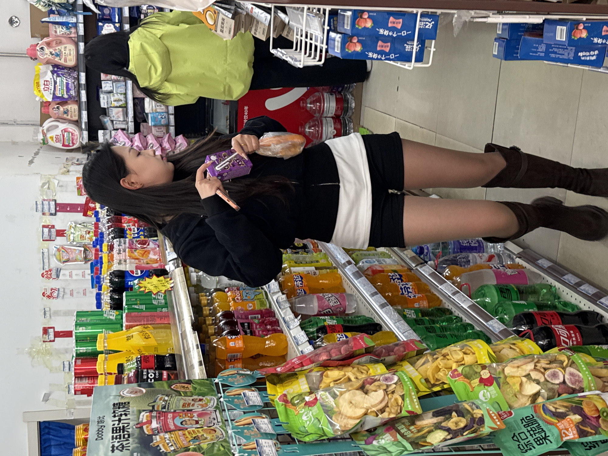 A young woman in Zhongtun, China, stands in a store aisle. She wears a black jacket, black shorts with a white band, and brown knee-high boots. She holds a purple package and a wrapped item. Shelves are filled with various products, including drinks, snacks, and cleaning supplies. Another person in a green jacket is visible in the background.