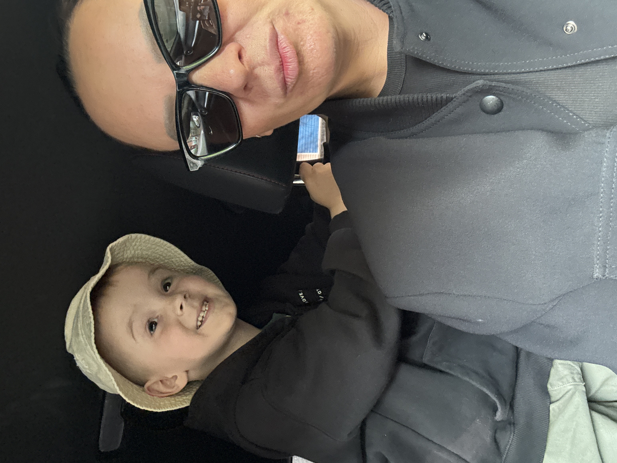 The image shows an adult wearing sunglasses and a dark jacket, positioned at an angle, possibly in a car, based on the black interior. A young child wearing a tan hat and black hoodie looks up at the camera. The child is holding a mobile phone. There is no information available in the image to identify the location as Chaikovsky, Russia.