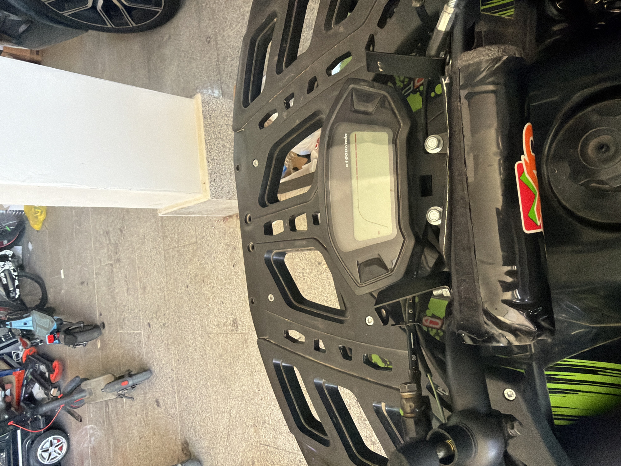 The image depicts the dashboard and front rack of a black ATV, along with the surrounding environment. The ATV's dashboard features a digital display indicating x1000r/min, suggesting it is a tachometer. A sticker is visible on the side of the ATV's components.
The background shows a concrete floor and a collection of bicycles and scooters, indicative of a storage area or garage. A white wall is also visible in the background.
There's no information available to pinpoint the location as Asenovgrad, Bulgaria.